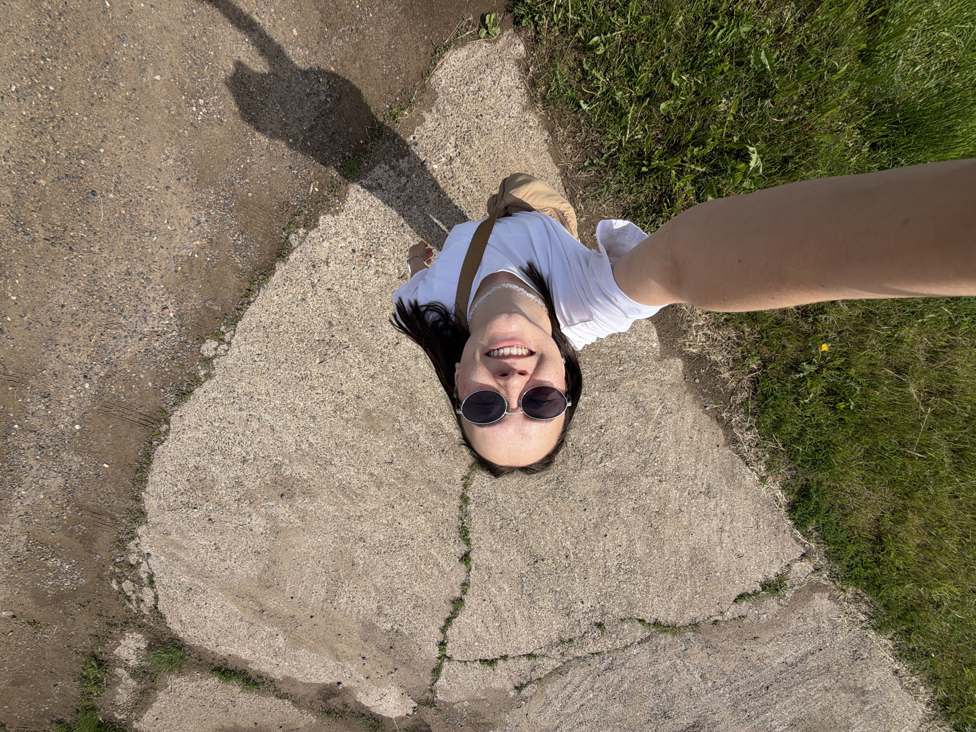 A young woman captures an overhead selfie outdoors in Lukintsy, Russia, on a bright and sunny day. The camera is held low, looking up at her from the ground. She is positioned on a cracked concrete surface that borders areas of dirt, gravel, and green grass.
The woman has dark hair and is wearing round, dark-lensed sunglasses, a white short-sleeved t-shirt, and light brown or khaki pants. A brown strap of a crossbody bag runs across her chest. She is smiling broadly, showing her teeth, and appears happy. Her face has visible freckles. Her right arm, bare, extends into the frame from the right side, holding the camera. A distinct shadow of her body, elongated by the sun, is cast on the ground to her left.
The ground features textured concrete with several prominent cracks where small patches of grass grow. This concrete transitions to gravelly dirt and lush green grass on the right side of the frame, with a small yellow flower visible in the grass. The bright lighting and strong shadow indicate clear weather during daylight hours. No visible text is present in the image.