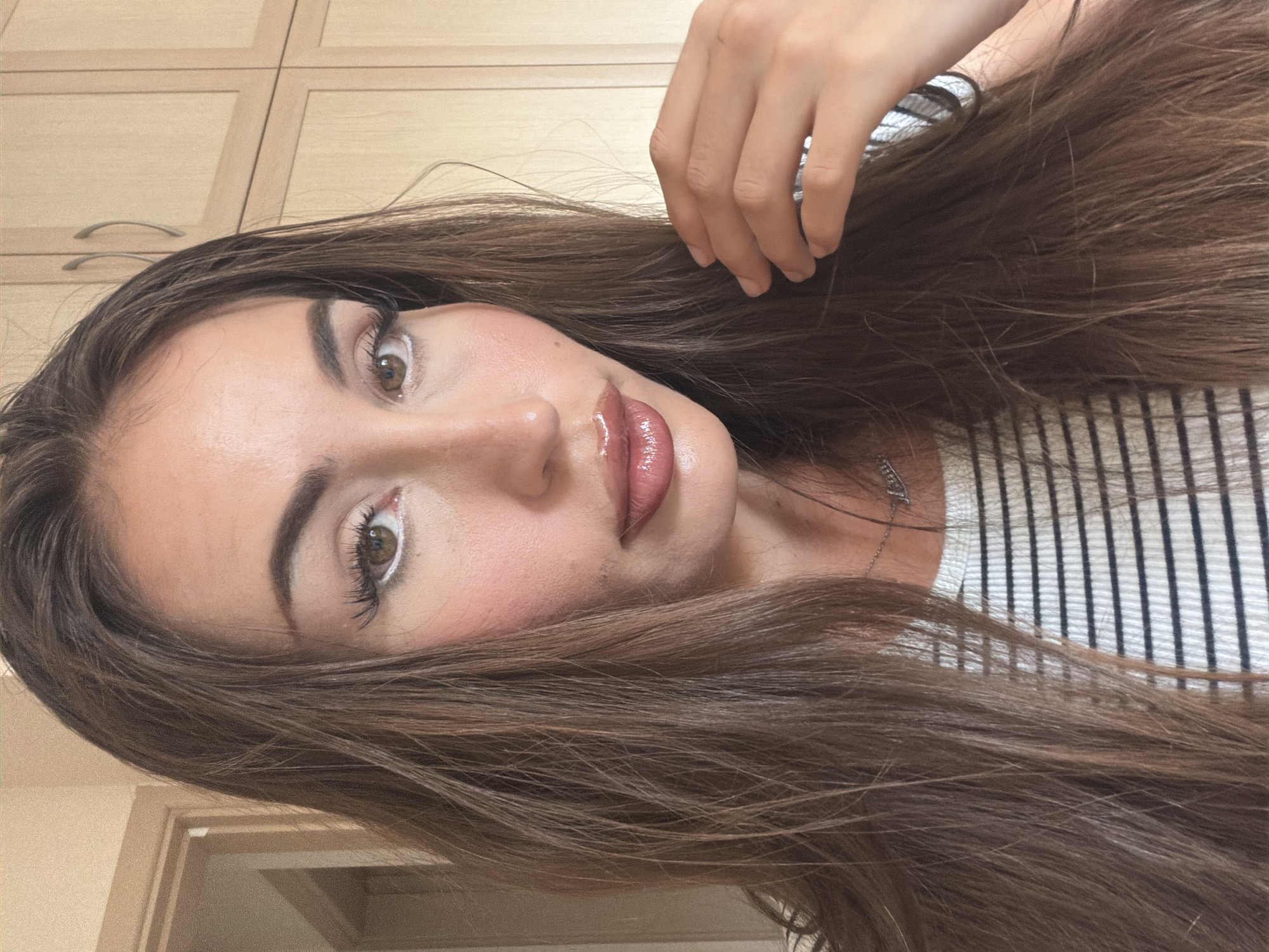 The image is a close-up, head-and-shoulders portrait of a young woman with long, brown hair. She is wearing a striped shirt and a necklace. Her makeup includes eyeliner, mascara, blush, and lip gloss. The background appears to be an indoor setting with wooden cabinets. There is no visible indication of the city of Tersefanou, Cyprus, in the image.