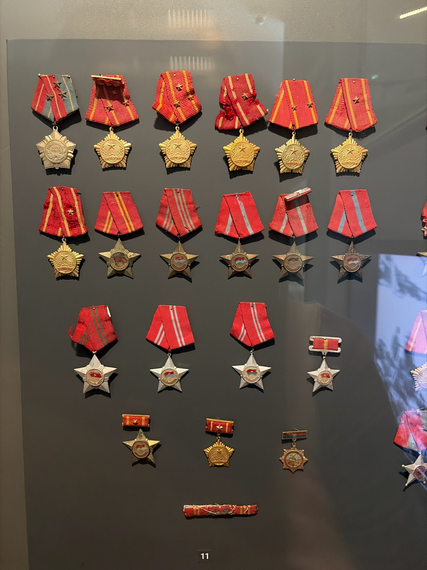 The image displays a collection of military medals, arranged in rows on a dark gray wall. The medals vary in design, with many featuring star shapes and red, white, and blue ribbons. Some medals have circular inserts with images or emblems. The arrangement suggests a museum exhibit or a private collection. The overall tone is formal and historical.
The image does not provide any information about its location in the city of Đà Nẵng, Vietnam.