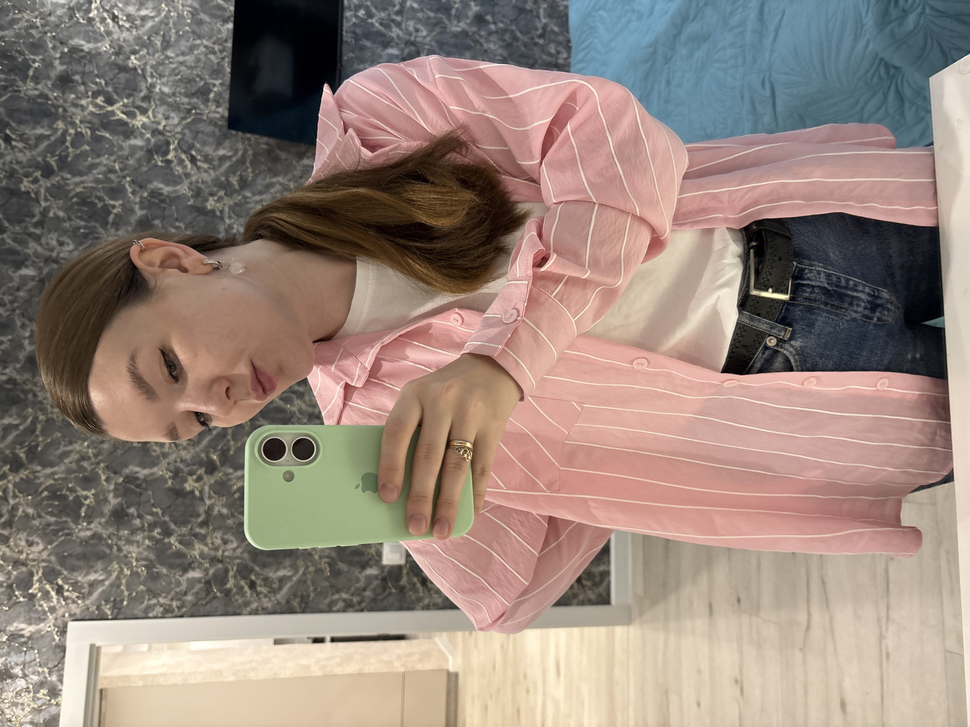 A young woman with light brown hair is depicted taking a selfie or mirror selfie indoors in Izhevsk, Russia. The image is oriented with the woman lying on her left side, but she appears to be standing or seated upright, holding a light green iPhone with the Apple logo visible on its case. She is looking towards the camera, maintaining a neutral and composed expression.
She is dressed in a casual yet stylish outfit, featuring a loose-fitting, long-sleeved pink button-up shirt with thin white vertical stripes. Beneath the pink shirt, a white t-shirt is visible. She wears blue denim jeans, secured with a black belt that has a silver buckle. Accessories include a gold-colored ring on her right ring finger and distinctive silver-toned cuff earrings with clear, possibly quartz, drop details that adorn her right ear.
The background elements suggest a modern indoor setting. A dark gray or black marbled wall covers the left side of the frame, providing a textured backdrop. To the right, a light blue textured fabric, possibly a blanket or bedspread, is visible. The lower part of the image shows a light wooden floor, and a white frame, possibly from a mirror or door, is also present. A dark, possibly black, object (like a screen or furniture piece) is partially visible in the upper left corner of the background. The overall lighting appears to be ambient indoor light.