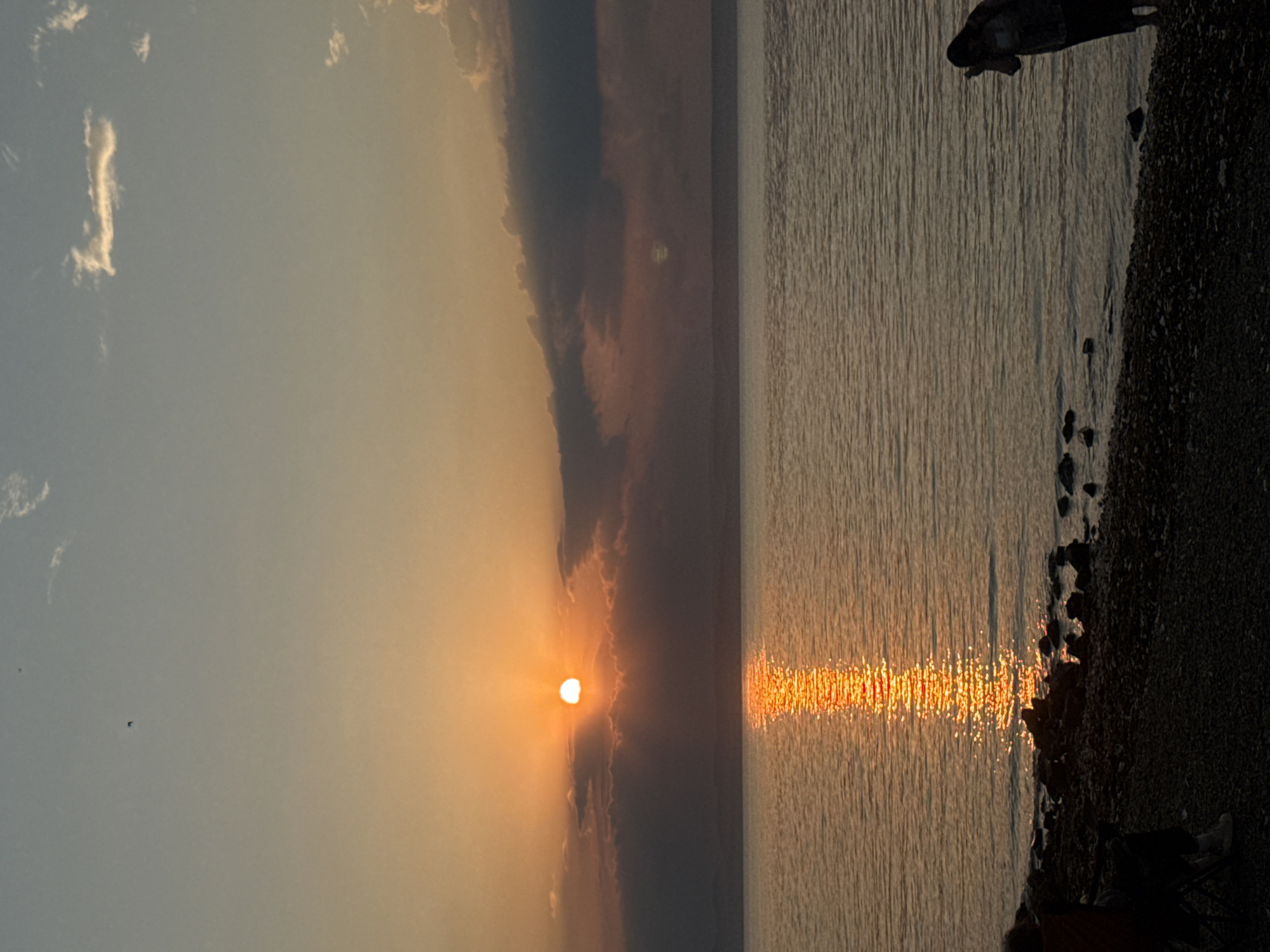 The image shows a sunset over a body of water, likely the sea given the location context of Vladivostok, Russia. The sun is low on the horizon, casting a warm, orange glow that reflects on the rippling water, creating a shimmering path of light. Clouds are present in the sky, some illuminated by the setting sun, and others darker and more dramatic. The foreground depicts a rocky shoreline with several dark stones scattered along the edge. In the upper right portion of the image, a dark, silhouetted object, possibly a person or a piece of equipment, is visible against the water. Further down the shoreline, a person is seated in a dark chair, also silhouetted, appearing to be enjoying the view. The overall atmosphere is serene and peaceful, indicative of a tranquil evening. The time of day is clearly sunset, and the weather appears clear with some cloud cover. The scene evokes a sense of calm and appreciation for nature's beauty. There is no visible text in the image.