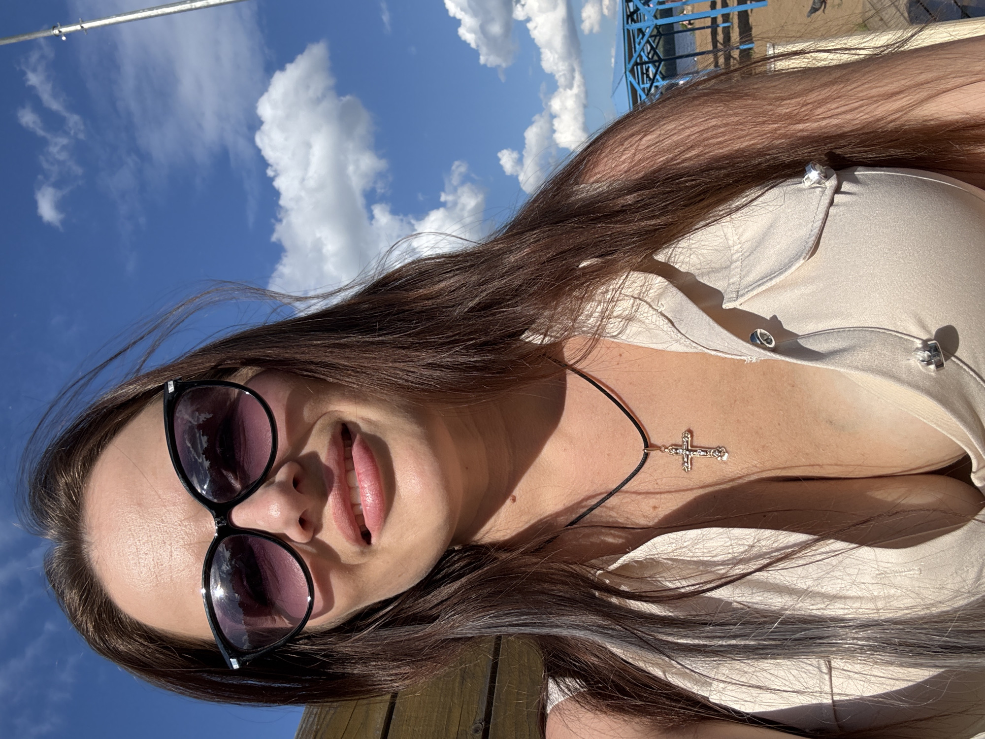 In a selfie taken outdoors, a woman with long brown hair and sunglasses smiles at the camera. She is wearing a light-colored, open-necked shirt with a crucifix necklace. The background shows a bright blue sky with scattered white clouds, a wooden structure, and a glimpse of a blue structure and water, suggesting a location near a body of water. The overall scene appears to be a sunny day. The location is the city of Chaikovsky, Russia.