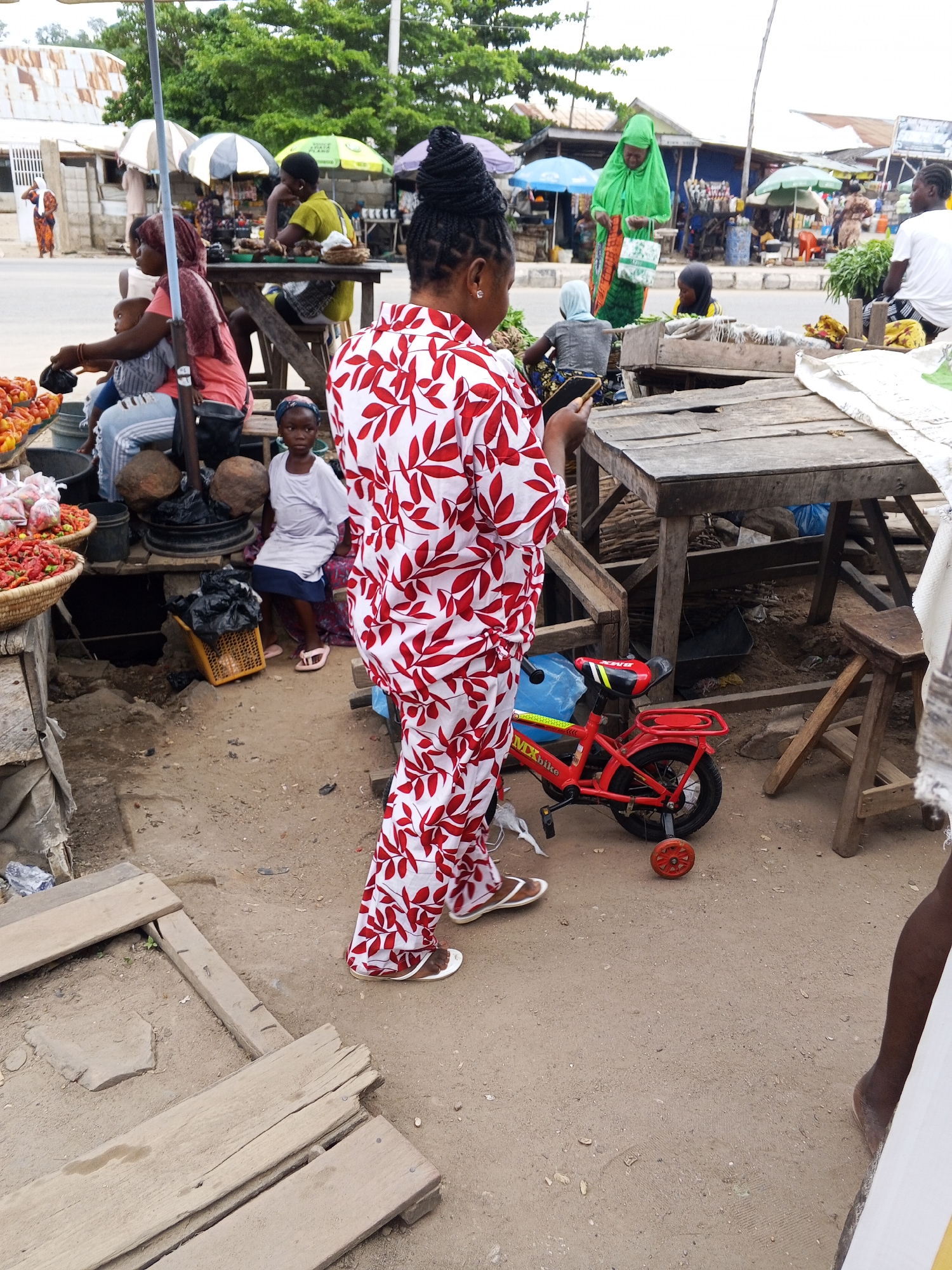 A group of people are gathered at an outdoor market in Zango-ndaji, Nigeria. The scene is bustling with activity, likely taking place during daylight hours. Several individuals, including women and children, are interacting with vendors and products. One woman in a red and white patterned outfit is holding a smartphone, while a young girl in a white top and blue skirt sits nearby. Another woman in a bright green hijab is browsing goods. Various market stalls are visible, displaying produce and other items. Wooden tables and benches are set up for vendors and customers. A small red children's bicycle with training wheels is parked on the dirt ground. The overall atmosphere suggests a lively and typical market day in the region.