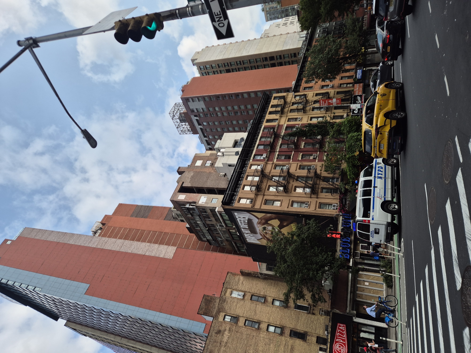 This is a street-level photograph taken in New York City, United States of America. The image captures a view looking up at several tall buildings, an intersection with traffic, and pedestrians. On the left side of the frame, a red neon sign for "PIZZA" is visible above a storefront. Next to it, a blue neon sign reads "BLUE POINT." A white NYPD police van is parked in the street, and a yellow taxi is in motion, along with other vehicles. A person is riding a blue bicycle in a bike lane. Pedestrians are seen walking on the sidewalk. The sky is partly cloudy. The buildings vary in architectural style and color, with some featuring brick facades and others modern glass exteriors. Traffic lights and street signs, including a "ONE WAY" sign, are present at the intersection. Fire escapes are visible on some of the older buildings.