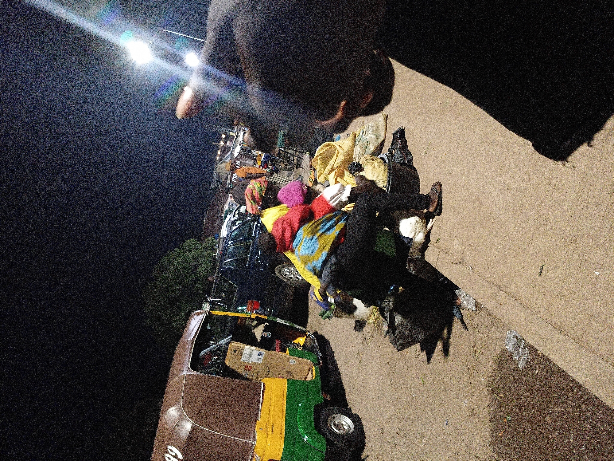The image depicts a nighttime scene in Bukuru, Nigeria, likely a roadside market or a similar public area. In the foreground, a person is crouched down, partially obscured by a large black shadow cast by the photographer's hand and arm. This person is wearing a bright, multi-colored jacket over a red top, dark pants, and patterned shoes. They appear to be interacting with or sorting through various items on the ground.
Beside the crouching person is a three-wheeled auto-rickshaw, painted green and yellow, with its door open. Inside the auto-rickshaw, several items are visible, including boxes and what appears to be cargo. Scattered around the auto-rickshaw and the crouching figure are several large, light-colored sacks, a dark bucket, and other assorted items, suggesting goods or belongings.
In the background, a dark-colored SUV is parked, and beyond that, more vehicles and what appear to be market stalls or structures are visible under artificial lighting. A bright streetlamp in the upper right corner of the image casts a strong glare and lens flare, indicating it is a significant light source in the scene. The ground is a mixture of dirt, gravel, and some areas of what looks like spilled liquid or debris. The overall impression is one of activity and commerce taking place after dark.