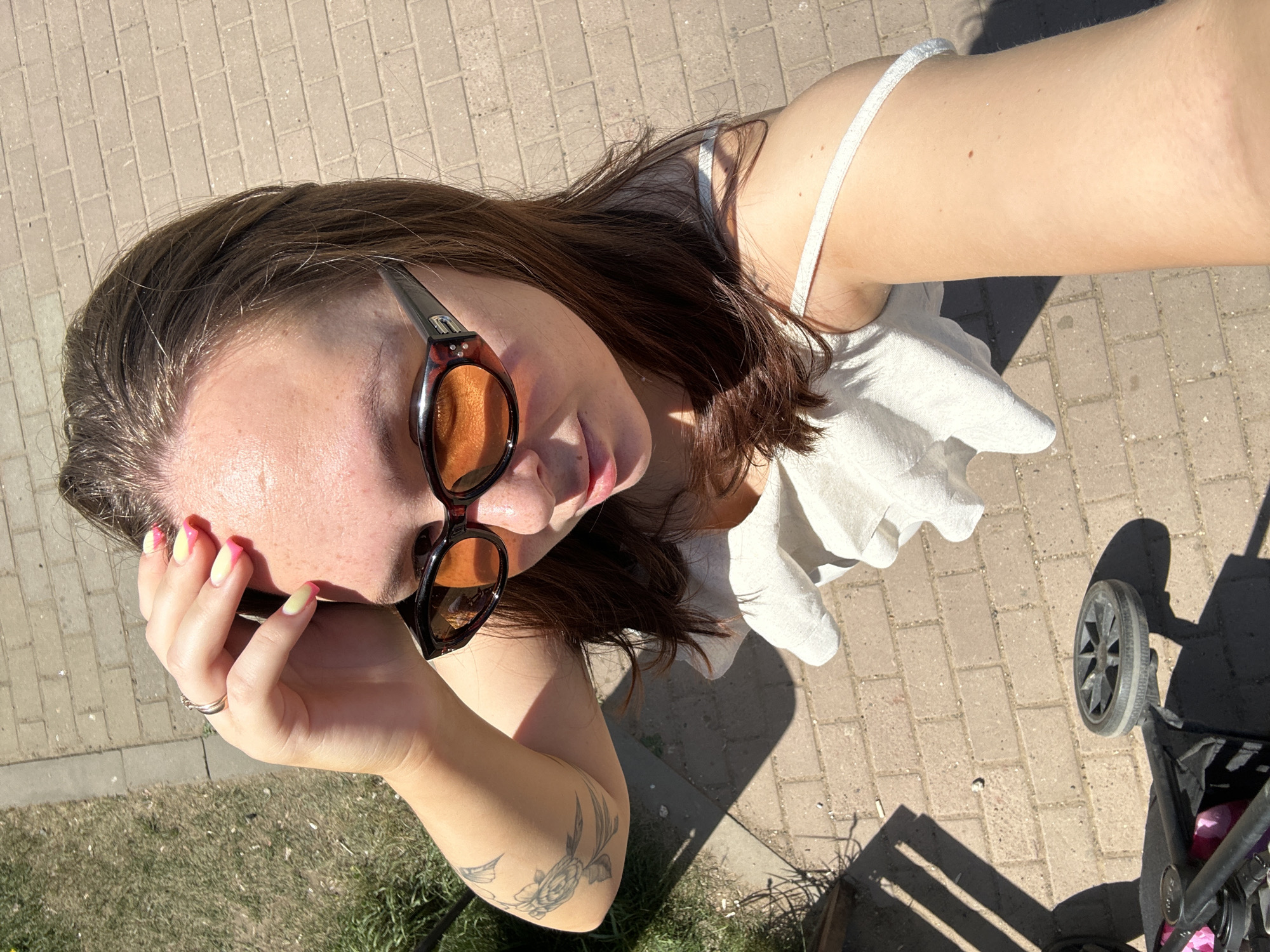 A woman in a white tank top is taking a selfie from a high angle. She is wearing sunglasses with amber lenses and has a tattoo on her left arm. She is holding her hand up to her face, showing a ring and nails with a two-tone pink and yellow design. To her right is a stroller on a brick pathway. Grass is visible along the edge of the walkway. The location is not explicitly identifiable as Chaikovsky, Russia, from the image.
