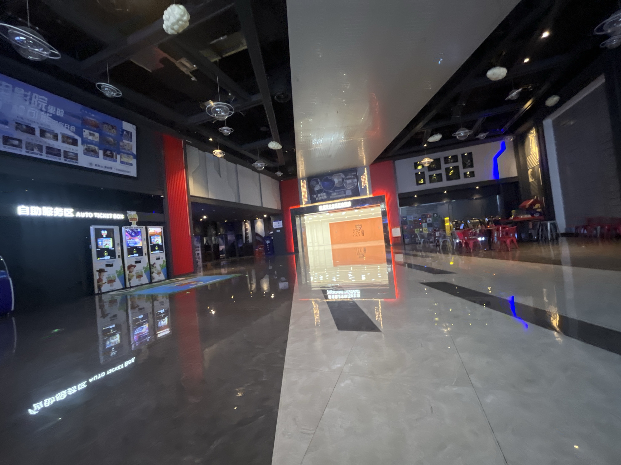 The image shows the interior of a cinema lobby in Shapingba District, China. The lobby features a polished floor that reflects the overhead lights and various elements of the space. On the left side of the image, there are several self-service ticket machines with colorful displays, some featuring characters from the movie Toy Story. Above these machines, a large banner with Chinese text and images of movie posters is displayed. The ceiling has a modern design with suspended light fixtures and decorative elements resembling planets.
Towards the center of the image, a large digital screen displays movie information. The screen is illuminated and appears to be showcasing trailers or promotional content. The surrounding area has red and white vertical elements, and there are some framed pictures on a wall in the background.
Further to the right, the lobby opens up to reveal seating areas with red chairs, suggesting a waiting area for moviegoers. The overall ambiance is typical of a modern cinema, with emphasis on lighting and visual displays to attract customers. The floor's reflectivity is a prominent feature, creating a sense of depth and highlighting the clean, contemporary design of the space.