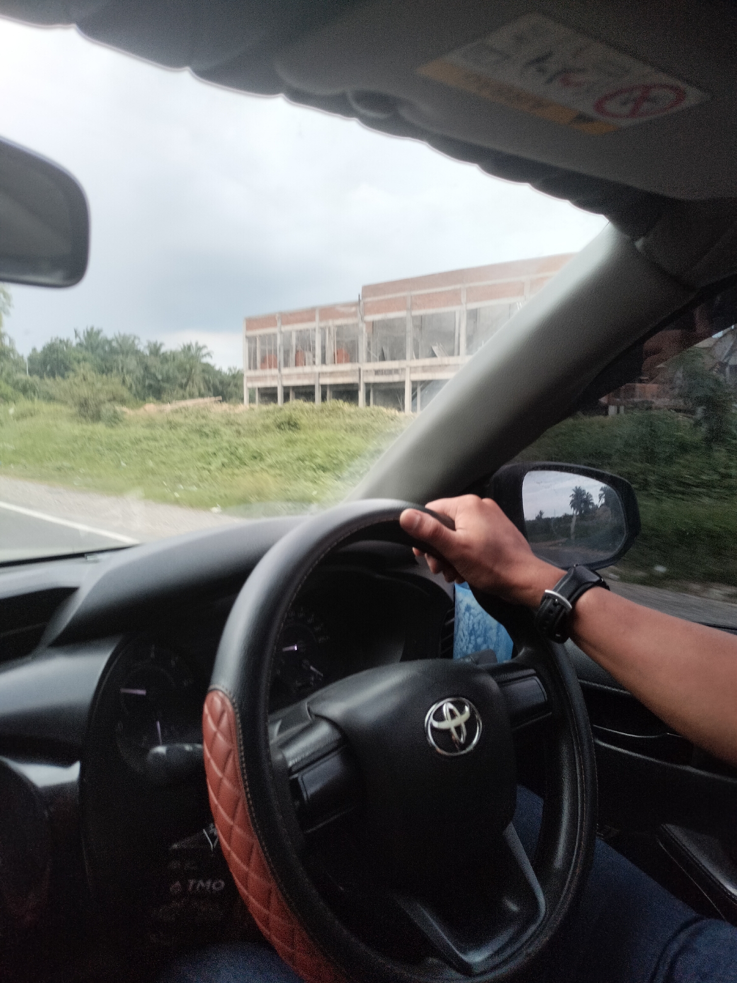 The image is a first-person view from inside a car, looking towards the steering wheel and out the windshield. The driver's hands are on the steering wheel, which has a brown diamond-stitched cover. A watch is visible on the driver's left wrist. In the foreground, the dashboard and part of the car's interior are visible. Through the windshield, a partially constructed building with an exposed brick facade and open window frames is seen in the middle ground. This building is surrounded by green vegetation, including what appear to be palm trees, suggesting a rural or semi-rural setting. The sky is overcast. The location is identified as Bukit Selamat, Indonesia, implying the scene is captured while driving in or near this area.