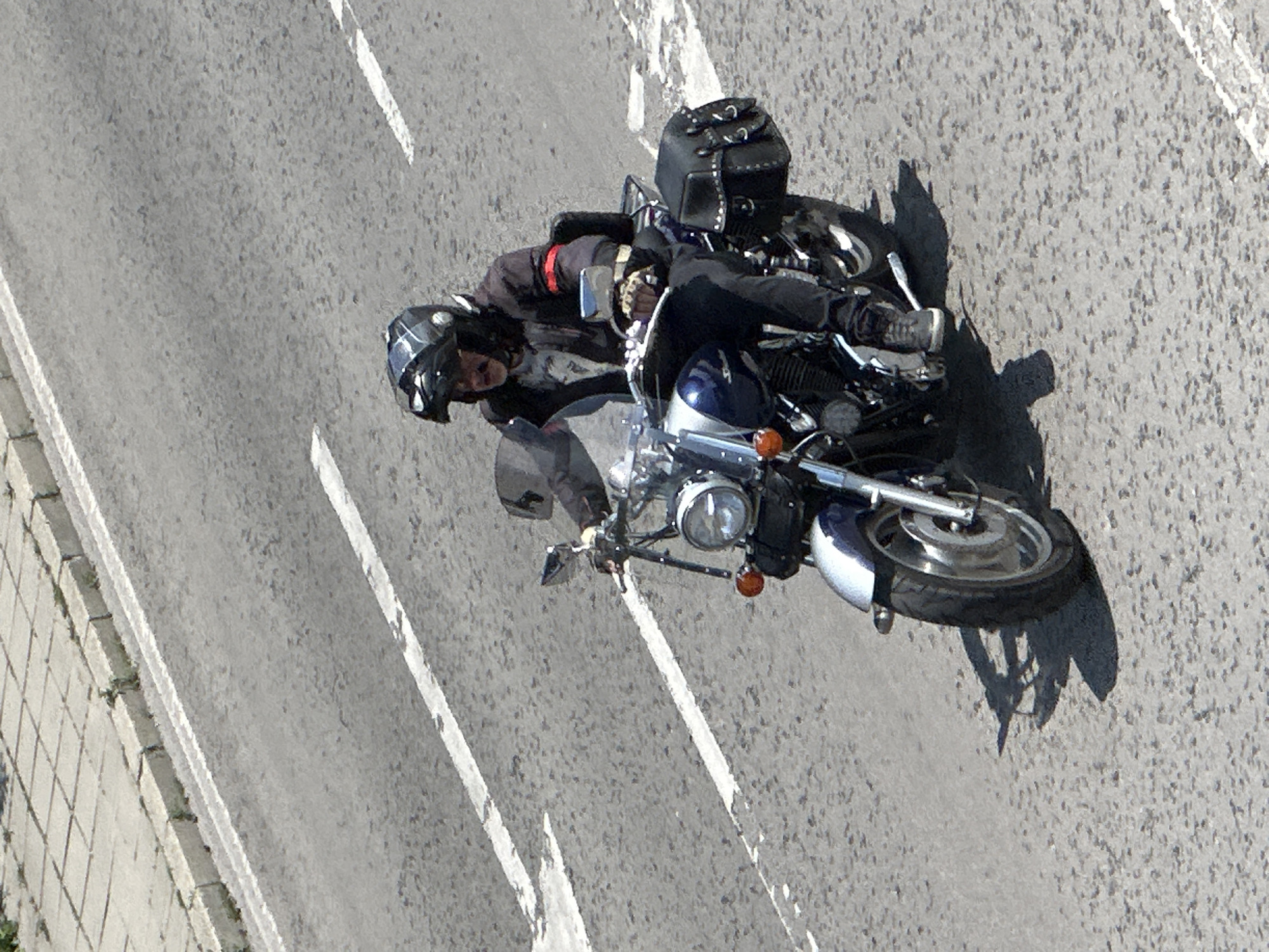 An overhead or high-angle shot captures a person riding a motorcycle on an asphalt road in Veliko Tarnovo, Bulgaria. The motorcyclist is wearing a dark full-face helmet, a dark jacket with subtle red accents on the arm, and dark pants. They are seated on a cruiser-style motorcycle, which features a prominent chrome headlight, a blue and silver front fender, and a black bag secured to the rear.
The road surface is gray asphalt, marked with white dashed and solid lines indicating lanes. A tiled sidewalk and curb run along the left side of the frame. Strong, distinct shadows of the motorcycle and rider are cast onto the road, indicating bright, sunny weather and likely midday or early afternoon sun. The motorcycle appears to be in motion or leaning into a turn. No discernible text is visible in the image.
