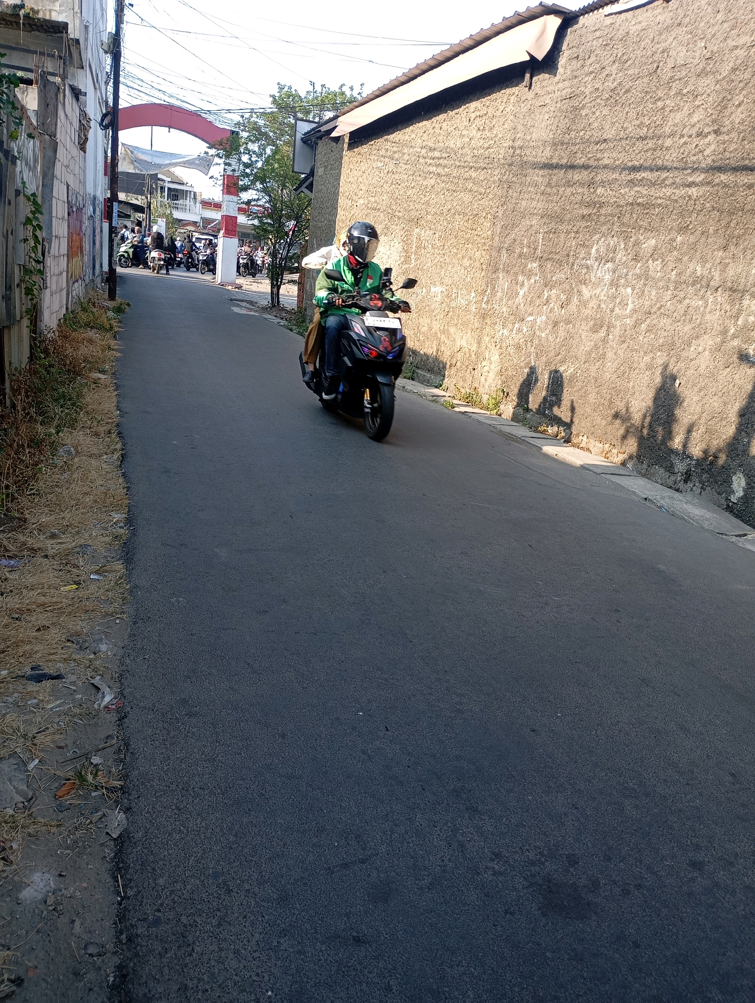 In a narrow asphalt street in Babelan, Indonesia, a person wearing a green jacket and a helmet rides a black scooter, casting a shadow on the textured concrete wall to their right. The wall runs alongside the street, with a corrugated metal roof extending over its top. On the left side of the street, a rough-textured building with exposed rebar and a graffiti-marked wall is visible. Further down the street, an archway with red and white striped pillars spans the road, with a banner hanging across it. Several other scooters and people are seen in the background, approaching the archway. The ground along the left side of the street is covered with dry grass and debris. The sky is clear and bright.