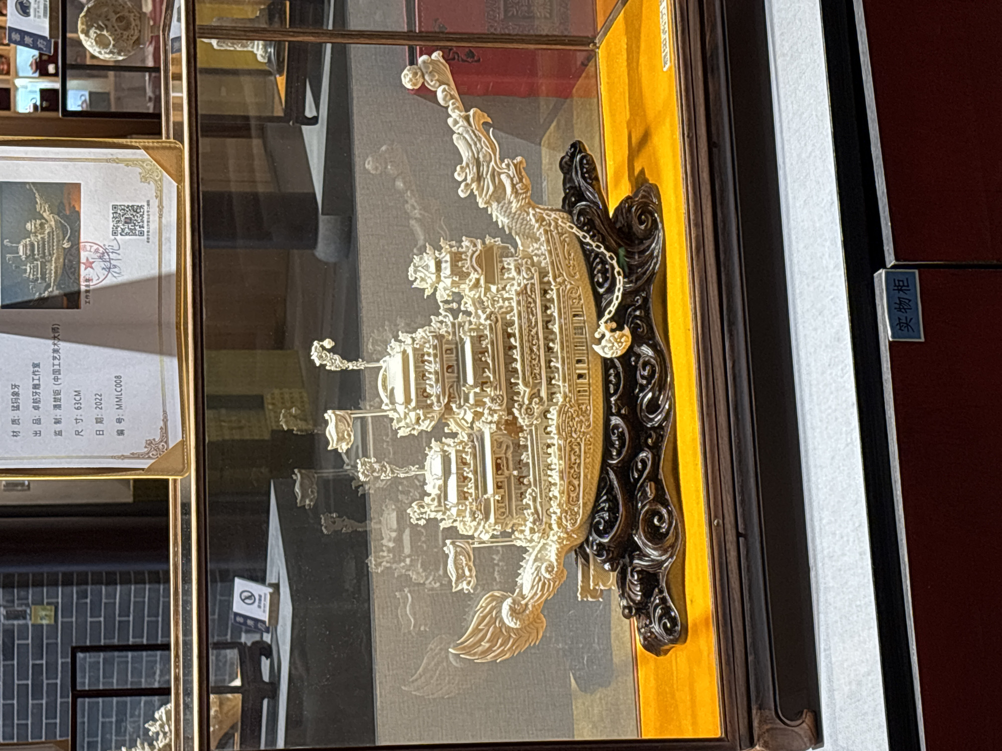The image shows a detailed ivory sculpture of a traditional Chinese dragon boat displayed in a glass case. The boat is mounted on an ornate dark-colored stand. A label is visible next to the sculpture, indicating it is made from mammoth ivory, produced by the Zhuofang Ivory Carving Studio, supervised by Pan Chuju (Chinese Arts and Crafts Master), measures 63 cm, and was created in 2022 with serial number MMLC008. The label includes Chinese text. The background shows a display cabinet, suggesting this is in a museum or gallery setting. The location in Khimki, Russia, is not evident from the image.