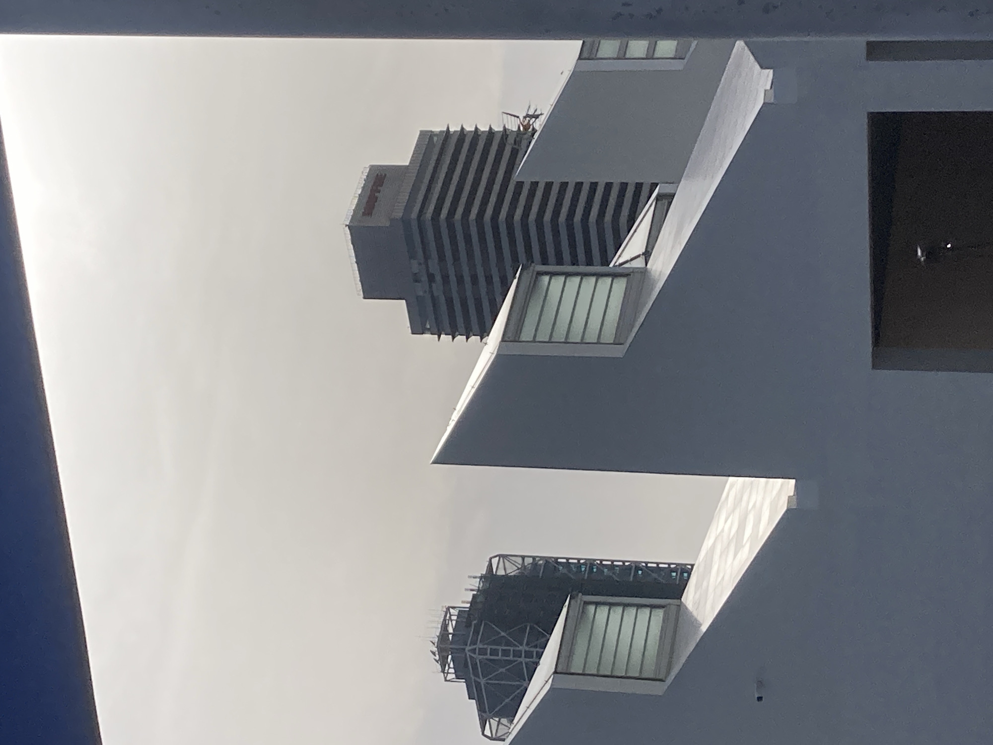The image shows an outdoor urban scene with a clear sky. In the foreground, there are the angular white structures of a modern building with slanted roofs and windows. Behind this, two tall buildings are visible against the sky. The building on the left appears to be under construction, with a visible scaffolding structure. The building on the right is a dark, rectangular skyscraper with horizontal bands and the red "MAPFRE" logo at its apex. This is likely the Mapfre Tower, a prominent skyscraper in Barcelona, Spain, located in the Port Olímpic area.