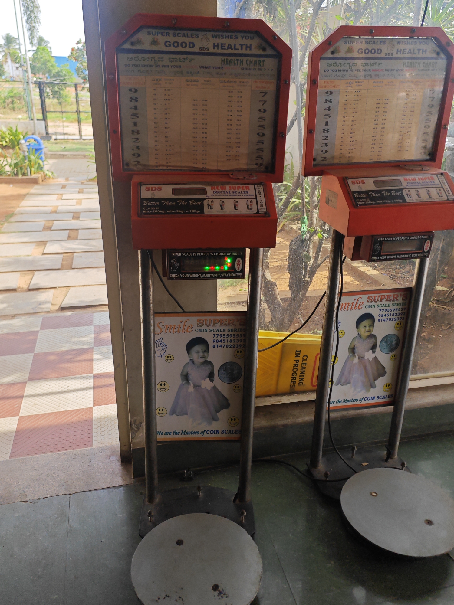 The image shows two coin-operated weighing scales, typical of those found in public places in India, positioned outdoors in what appears to be the city of Aruru. The scales are red and feature health charts and digital displays. One scale has a sign that reads "Smile SUPER'S COIN SCALE SERIES" and a picture of a baby. The background shows some trees and foliage, suggesting a somewhat rural or semi-urban setting. The ground is paved with tiles.