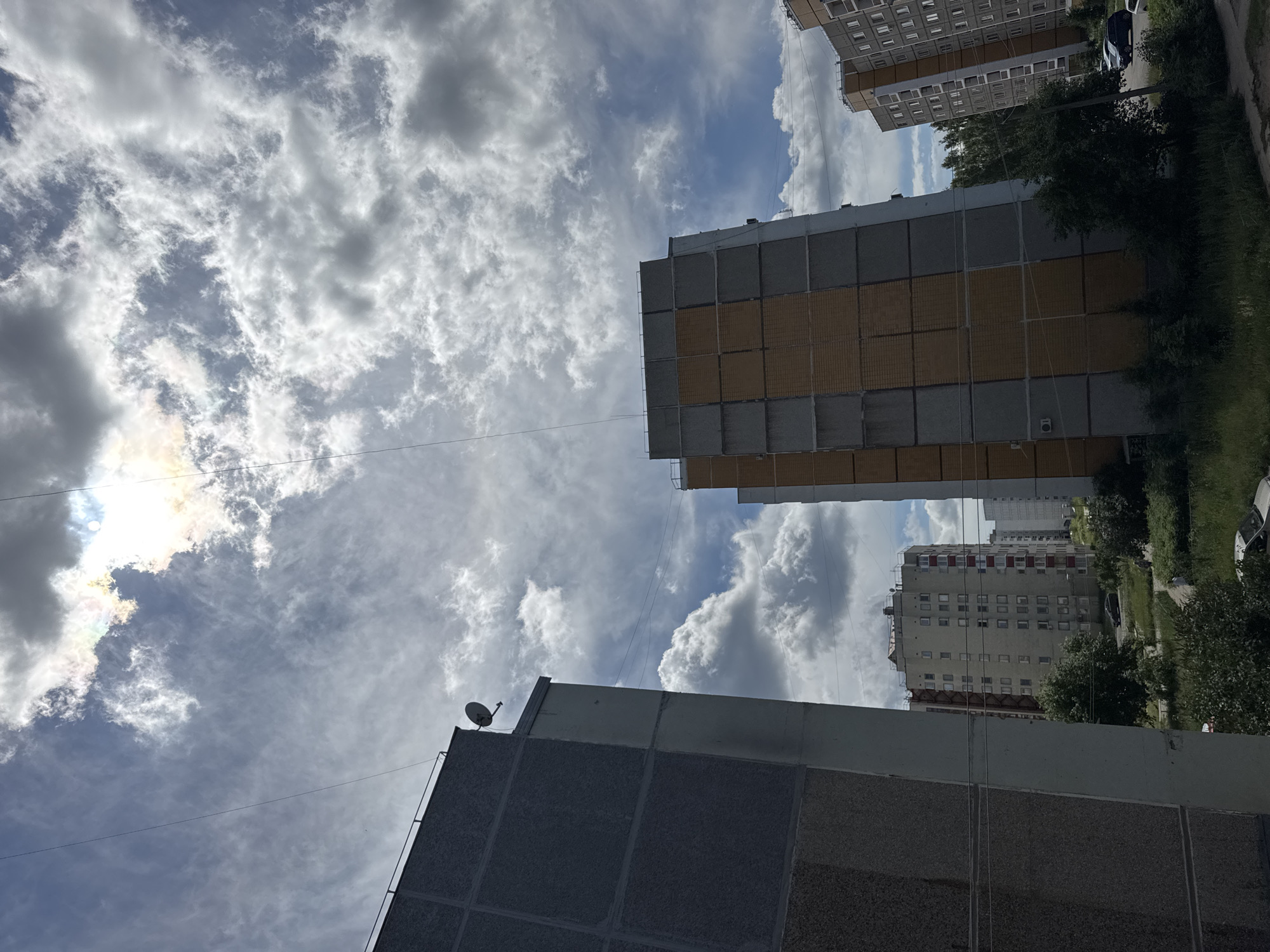 The image shows a view of apartment buildings in the city of Chaikovsky, Russia, under a cloudy sky. The sky is partially obscured by clouds, with patches of blue sky visible and the sun creating a bright halo effect. In the foreground on the left is the side of a tall, grey apartment building with a rough texture and some concrete sections. To the right of this building, and slightly further back, is another taller apartment building with a distinctive alternating pattern of grey and yellow-orange facade panels. Further in the distance, more apartment buildings are visible, appearing lighter in color. Green trees and vegetation are present at the base of the buildings and in the surrounding area, with a patch of grass and a dirt path visible in the lower part of the image. A few cars are parked on a road or lot in the mid-ground. There are no people clearly visible in the image. Overhead, thin wires are stretched across the frame, likely utility lines.