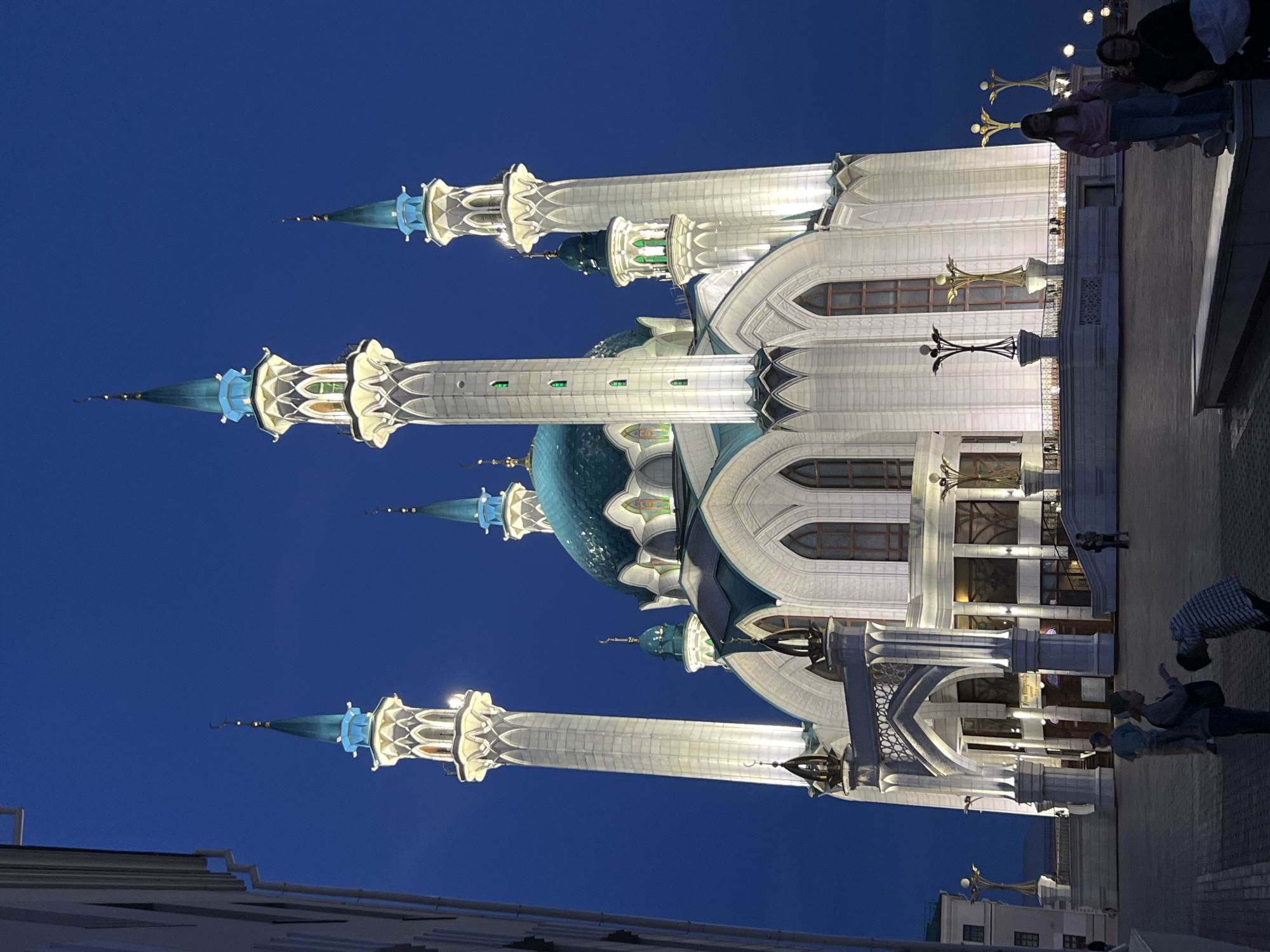 The image captures the Kul Sharif Mosque in Kazan, Russia, at night. The mosque's architectural features, including its minarets and central dome, are illuminated against a deep blue sky. People are visible on the right side of the image, on the pavement in front of the mosque. Some stand and interact, while others appear to be in motion. The area around the mosque is lit, with streetlights and building lights adding to the overall brightness of the scene. Buildings are visible at the bottom.
