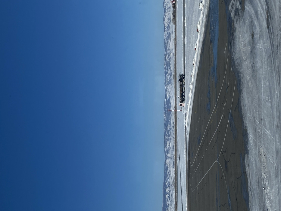 The image shows a bright blue sky dominating the left half of the frame. On the right is an outdoor, snow-covered area with mountains in the distance. A dark-colored train is visible on a straight track near the mountain range. The ground, partially covered in snow and ice, shows marked lines and dark patches of what appears to be a road or runway.
