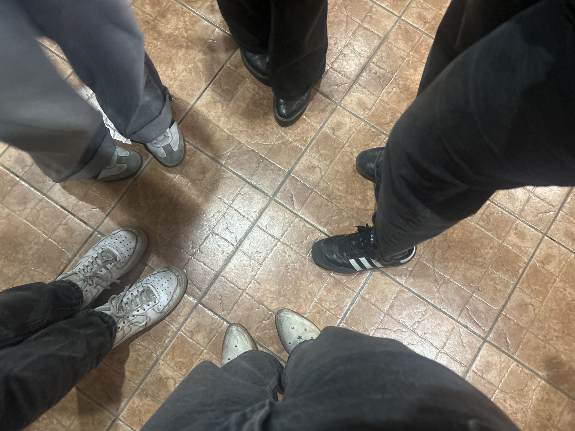 The image shows a view of the lower bodies of five people standing in a circle on a tiled floor. Each person is wearing different footwear: grey suede shoes, black leather shoes, black Adidas sneakers, white sneakers, and grey suede cowboy boots. The people are wearing trousers of different colors, including grey, black, and denim. The tiled floor appears to be inside a building. The image location is San Juan Cuautlancingo, Mexico.