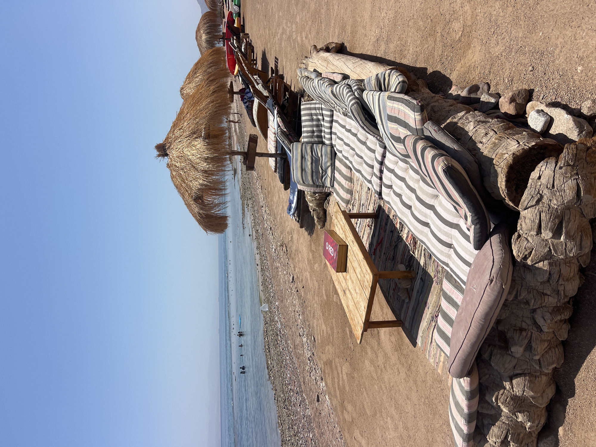 The image shows a beach scene in Nueiba, Egypt. In the foreground, a rustic lounge area is set up on a large log base, adorned with striped cushions. Adjacent to this seating area is a wooden table with a red book on it, positioned on a striped rug. Behind this, a row of sun loungers and thatched umbrellas are arranged on the sandy beach. The sea is visible in the background, with several people swimming and a few boats or kayaks in the water. The sky is clear and blue, and the overall environment suggests a relaxed, sunny day at the beach.