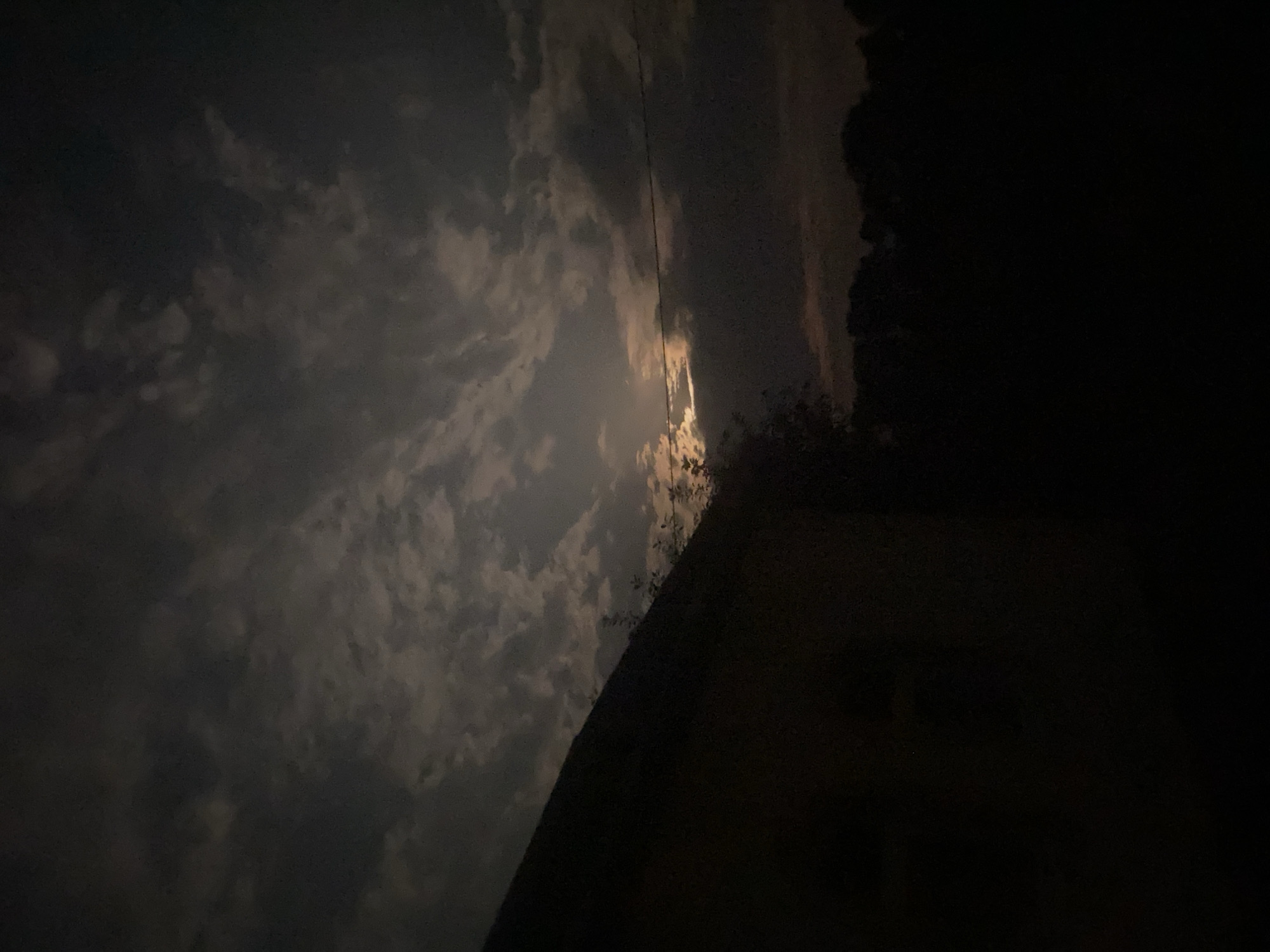 A nighttime image shows a dark silhouette of a house or building on the left side, with its roofline sloping down towards the right. The sky above is filled with clouds illuminated by a hazy moon, creating a dramatic and moody atmosphere. In the background, a dark line of trees can be seen silhouetted against the faintly lit sky. A single power line stretches horizontally across the middle right of the frame. The image appears to have been taken outdoors in Zastavna, Ukraine, during the night.