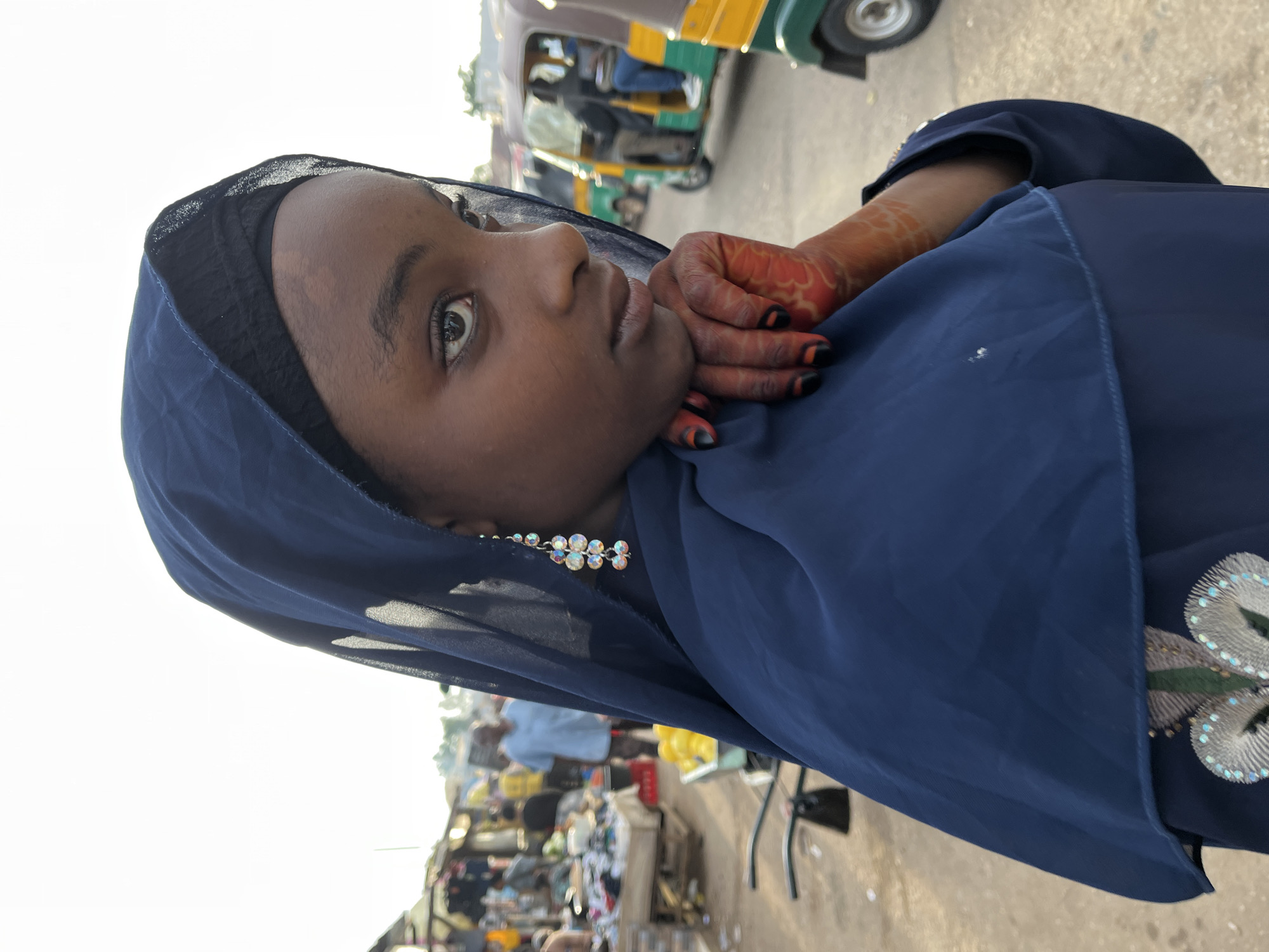 A young Nigerian girl, wearing a navy blue hijab and matching dress, stands in a bustling marketplace in Gura Top, Nigeria. Her hand rests on her chin, adorned with henna designs, and a sparkly earring dangles from her ear. In the background, market stalls are visible, filled with various goods, and colorful auto-rickshaws navigate the crowded street. The sun is setting, casting a warm glow over the scene.