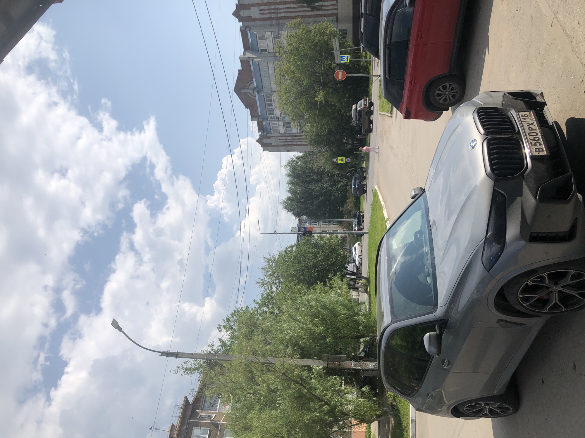 An eye-level, medium shot captures a street scene in Solikamsk, Russia, under a partly cloudy sky. In the foreground, a grey BMW X6 is parked on asphalt. To the right, a red SUV is parked alongside it. Beyond these vehicles, a street curves towards a residential building with an ornate facade. Trees line the sidewalk, and a street lamp stands on the left. A woman in a pink shirt and white pants is walking across the street, with a dark SUV and a light-colored car visible in the background. Traffic signs and power lines are present throughout the scene.