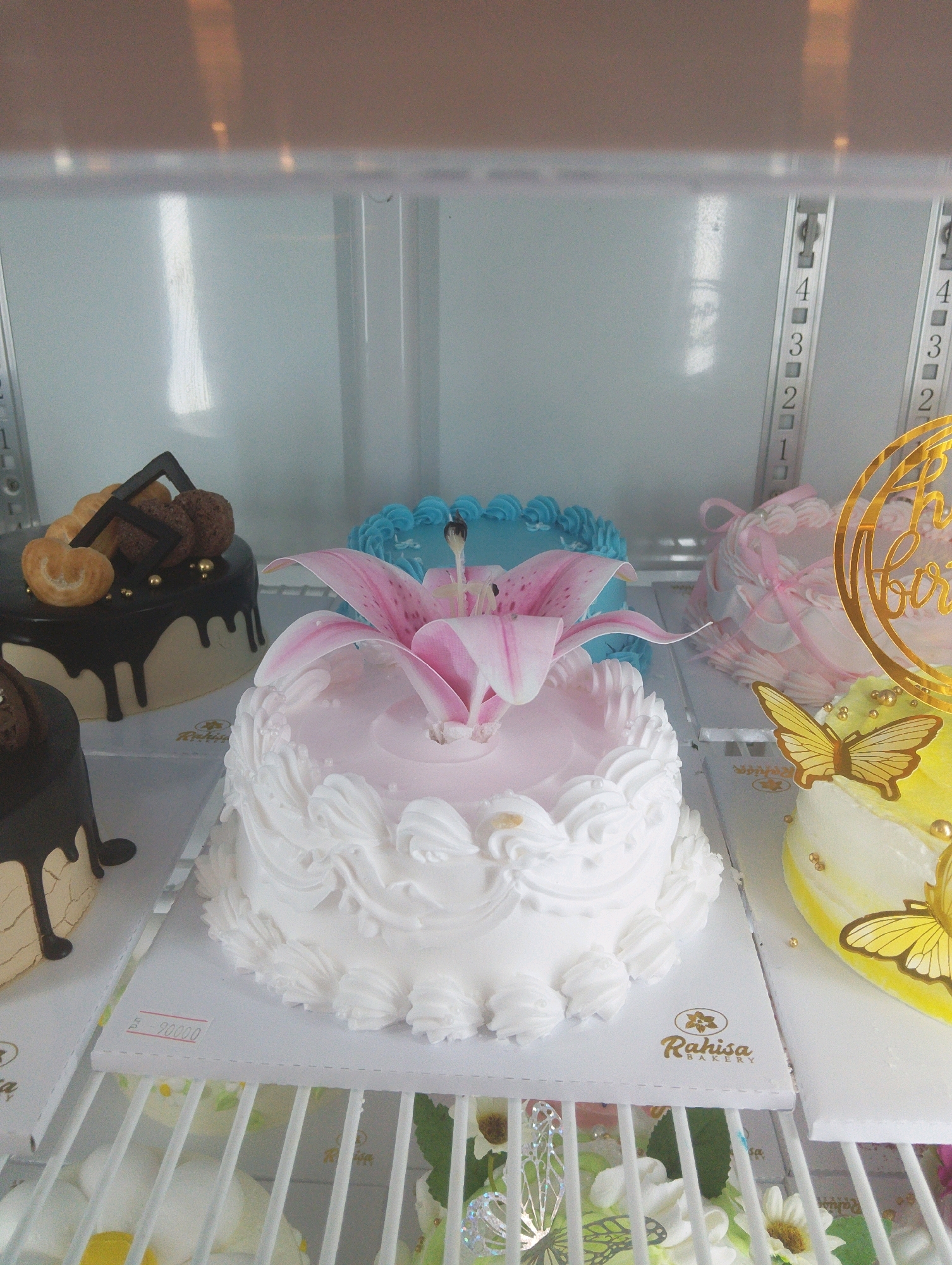 This is a photograph of cakes displayed in a refrigerated case, likely in a bakery. The lighting suggests the case is illuminated from above, casting reflections on the glass.
In the foreground, a large, round cake is decorated with white frosting piped in a swirling, floral pattern around the sides. The top of this cake is light pink and features a large, delicate pink lily as a centerpiece, with its petals unfurling outwards. To the left and slightly behind this cake is a dark chocolate cake with a drip glaze effect and topped with chocolate decorations and what appear to be small, golden spheres. Further back and to the left, a portion of another cake is visible, also with a dark chocolate drip glaze.
To the right of the main pink and white cake, a bright yellow cake is partially visible, adorned with golden edible beads and two golden butterfly decorations. Behind the pink and white cake and to the right, a pale pink cake is visible, decorated with ribbons and pearls. To the left of the yellow cake, a vibrant blue cake is seen, also with frosting decorations.
The cakes are arranged on shelves within the refrigerated case. In the background, vertical metallic strips with numbered markings (1, 2, 3, 4) are visible, possibly indicating shelf levels or temperature control. The overall impression is of a well-stocked bakery display. The brand name "Rahisa BAKERY" is visible on a white card beneath the pink and white cake. The presence of price information "-90000" on a sticker on the white card suggests this is a retail environment. The context implies this scene is located within Manaruwi, Indonesia, as specified.