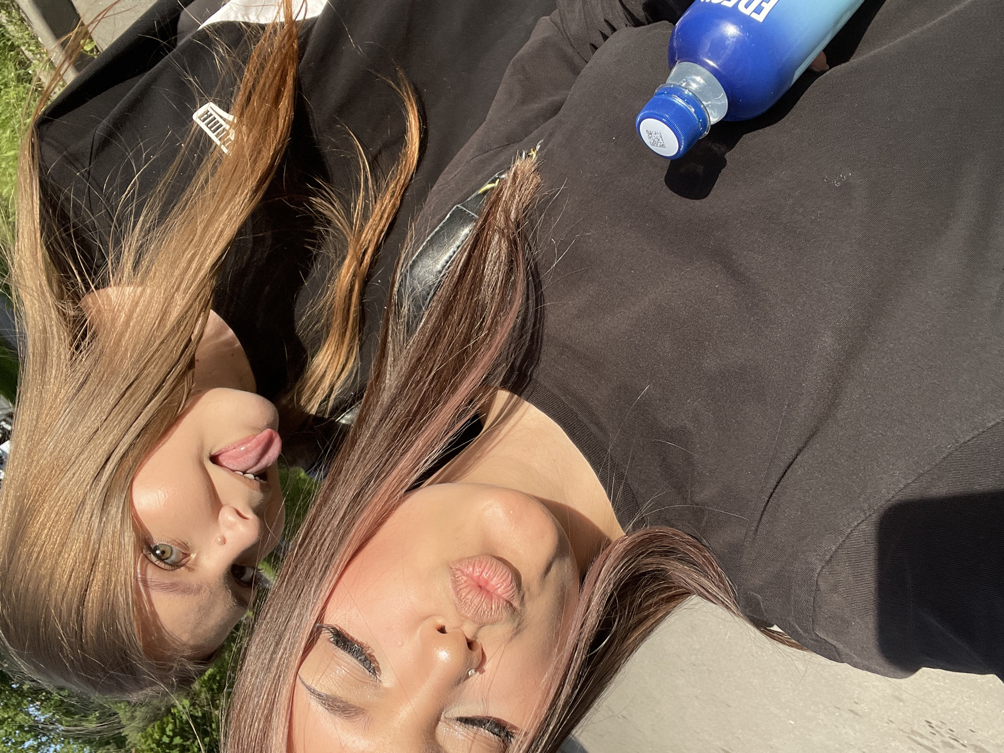 The image displays two young women in a tilted, close-up outdoor selfie, presumably taken in Saint Petersburg, Russia, under bright natural sunlight.
In the foreground, on the left, is a woman with long, dark brown hair subtly highlighted with reddish tones. She is seen from the mid-chest upwards, with her eyes closed and her lips puckered into a "kiss" pose. A small silver nose ring is visible on her left nostril, and a faint silver hoop earring is on her right ear. She wears a plain black long-sleeved top, and a black strap, likely from a backpack or bag, is visible over her right shoulder. Her skin is well-lit, showing highlights on her forehead, nose, and lips.
Behind and above her, on the right side of the frame, is a second woman with long, light brown/caramel-colored hair. She is smiling playfully with her tongue extended outwards. Her eyes, appearing greenish-brown, are open and directed towards the camera. A small, light-colored blemish or mole is located above her upper lip. She also wears a black long-sleeved top, distinctly featuring a white PUMA logo on the left chest area, partially covered by her hair.
In the bottom right corner, a blue and light blue gradient plastic bottle with a blue screw cap, featuring a white QR code sticker, rests on or near the foreground woman's chest. The word "FRESH" is partially visible on the bottle.
The environment includes a light-colored, possibly concrete or stone, paved surface in the bottom left. The background consists of green foliage, including trees and grass, indicating an outdoor, park-like or urban green space setting. Strong natural sunlight illuminates the subjects, creating distinct highlights on their skin and hair.