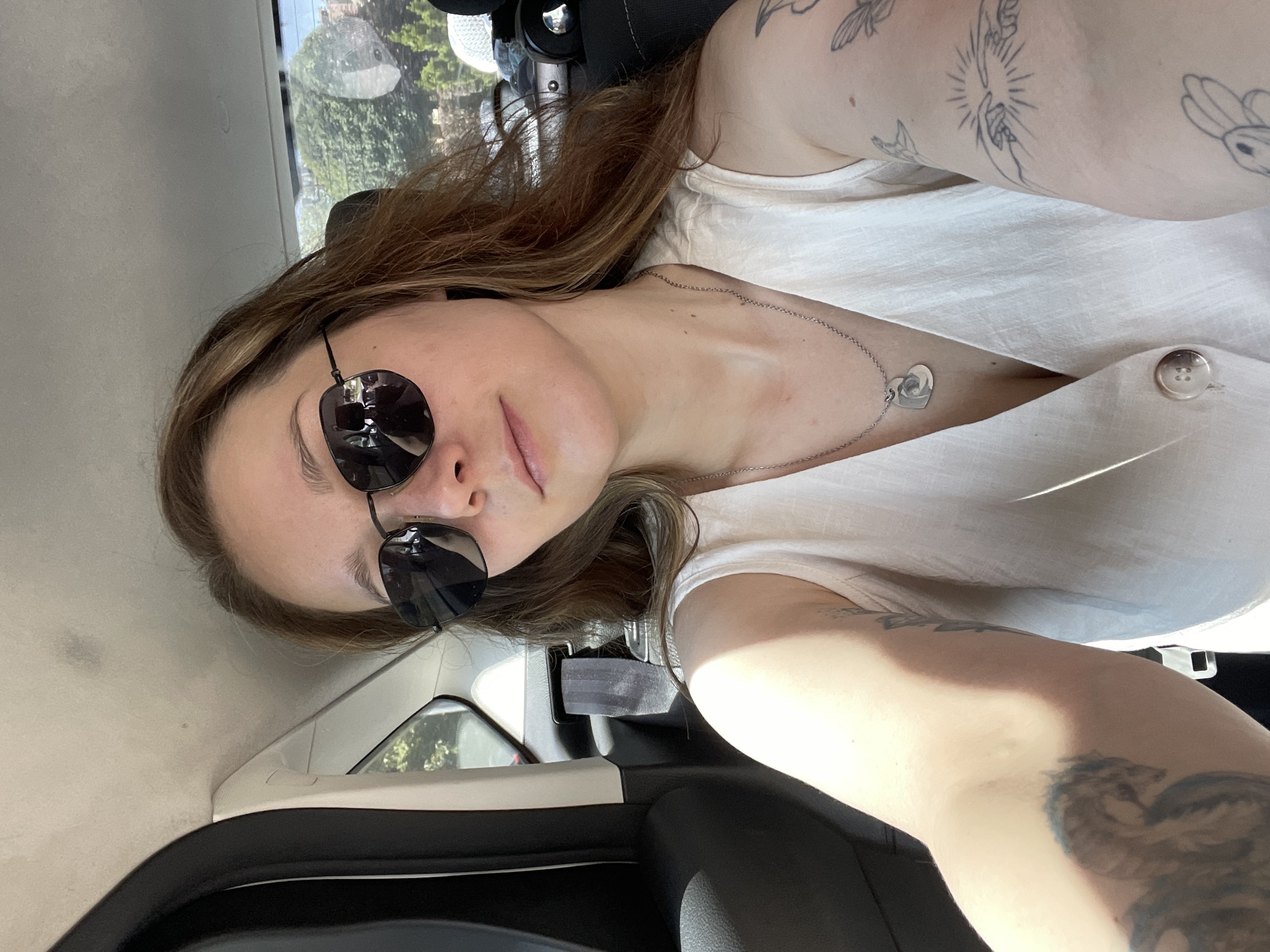 A woman with light brown hair and sunglasses is taking a selfie in the back seat of a car. She is wearing a white sleeveless dress with a V-neck and a silver necklace with a heart-shaped pendant. She has tattoos on both arms, including a dragon on her left arm and a depiction of the creation of Adam on her right arm. The interior of the car is dark, with a black seat and a visible seatbelt. Outside the car window, trees and buildings are visible, suggesting an urban or suburban setting, possibly in Stavropol, Russia. The lighting indicates it is daytime, with sunlight casting shadows on the woman's arms and face.
