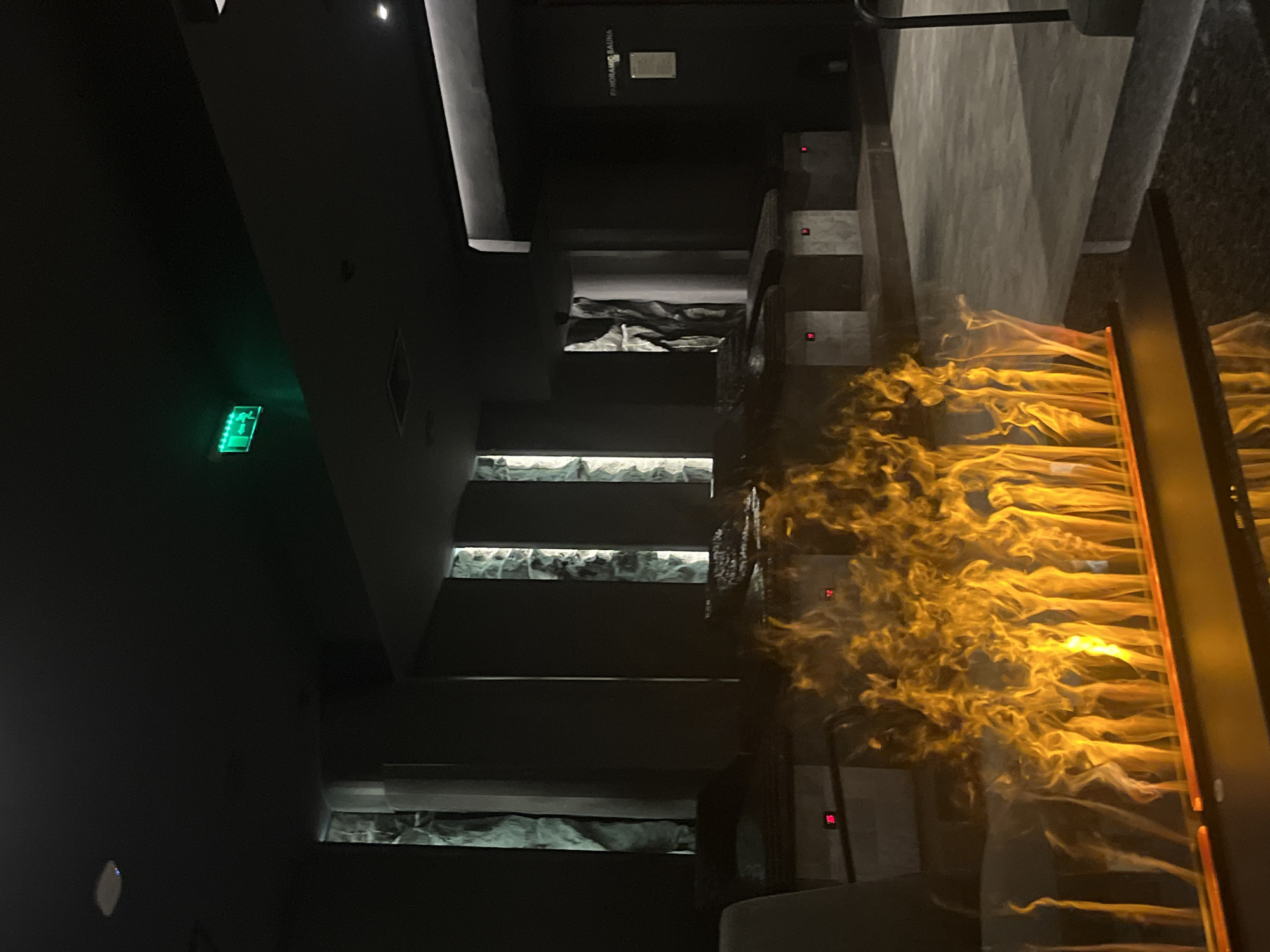 The image shows the interior of a building in Sofia, Bulgaria. A set of stairs with embedded lighting and decorative rocks is visible. A fire element, possibly a fireplace or artistic installation featuring flames, is present to the right of the stairs. An exit sign is visible on the wall on the left. The lighting is dim and dramatic. A sign reads "Panoramic Sauna."