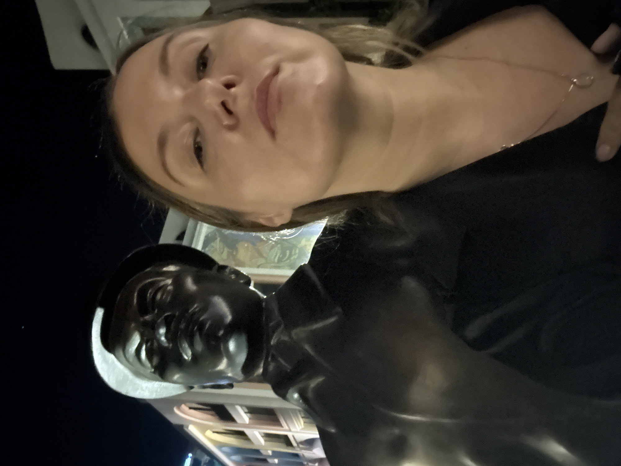 A woman is standing next to a statue of a sailor in the Bac Natural Heritage Area Trang, Vietnam. The woman has blonde hair and is wearing a black top and a necklace. The statue is of a man in a sailor's uniform with a white hat. The background shows buildings and a dark sky.