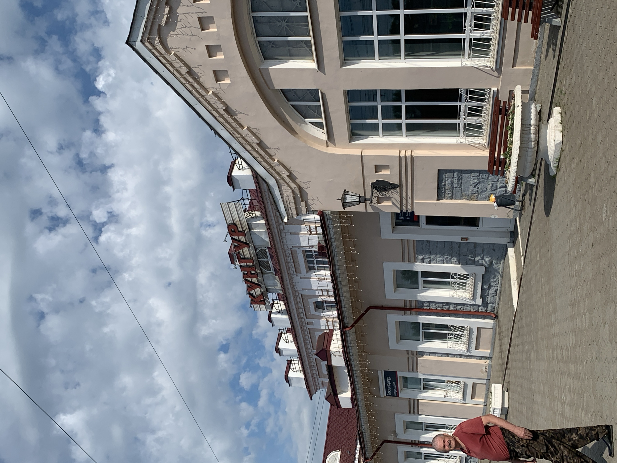 The image shows a man lying on the ground in front of a building in Kungur, Russia. The man is wearing a red t-shirt and camouflage pants. He appears to be relaxed and is looking at the camera. The building behind him is light beige with several windows and decorative trim. A sign with Cyrillic text is visible on the building. The sky is partly cloudy. The overall impression is of a casual outdoor scene on a bright day.