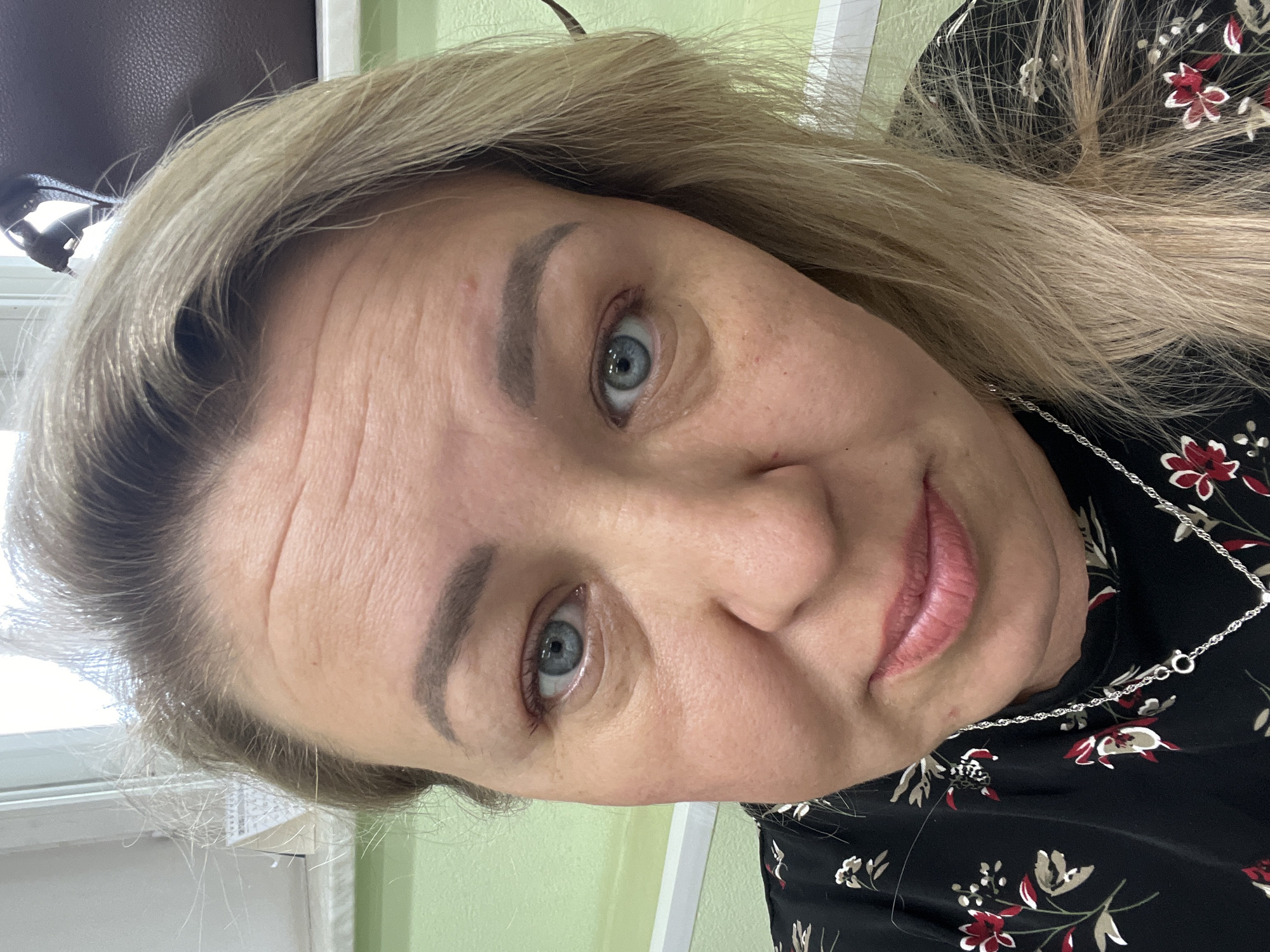 A close-up portrait of a middle-aged woman with blonde hair and blue eyes, wearing a black floral print top and a silver chain necklace. Her face shows wrinkles, and her eyebrows are lightly filled in. The background is a light green wall with a partial view of a window frame. The location is unknown. There is no indication in the image that this is in Chaikovsky, Russia.