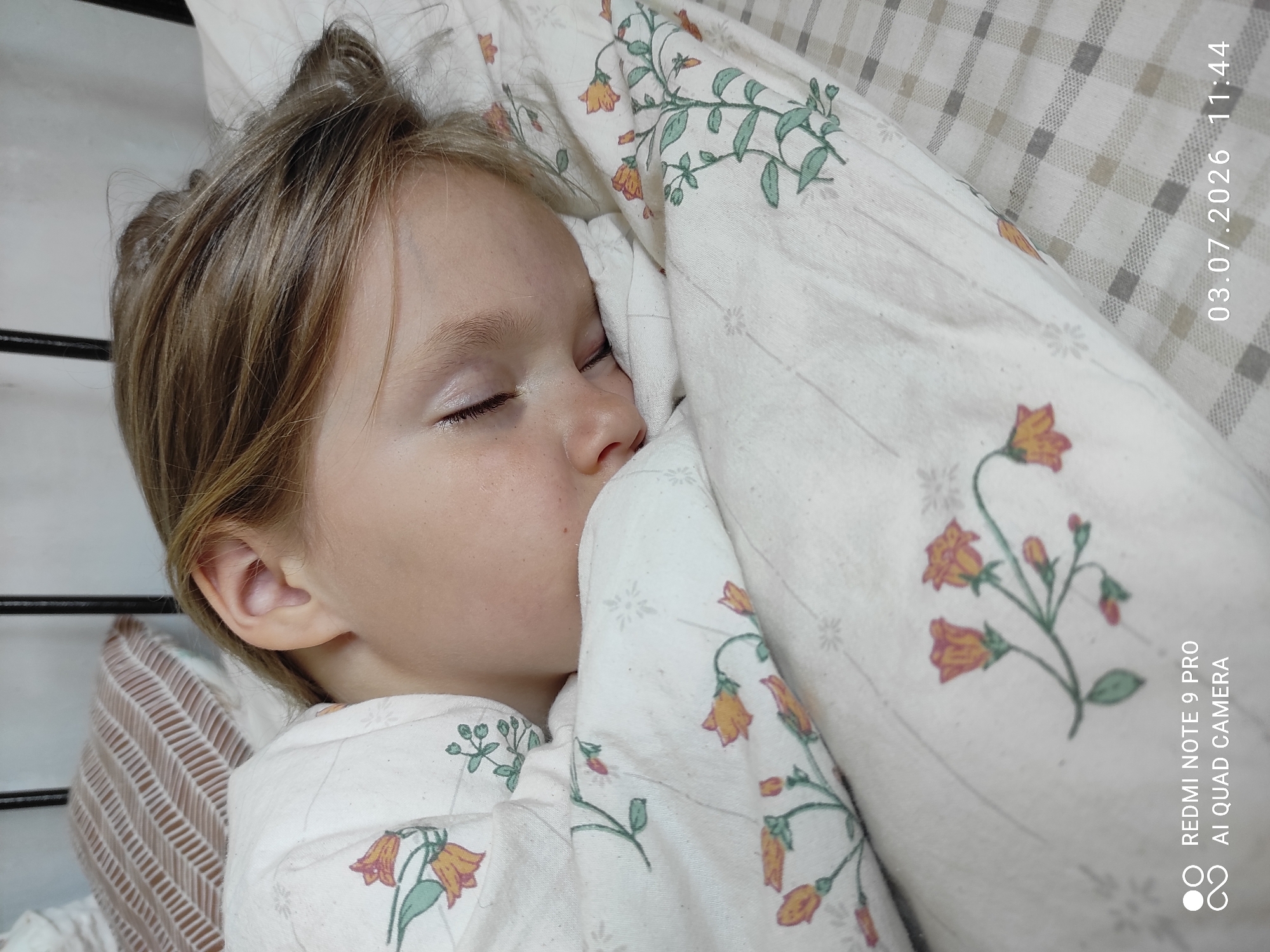 The image shows a close-up, eye-level shot of a young child sleeping in a bed. The child's head is turned to the right, with their eyes closed and hair resting on a patterned pillow. The child is covered with a white duvet featuring an orange and green floral pattern. The background shows a portion of a bed frame and another pillow with a striped pattern. A timestamp in the lower right corner indicates the date and time as 03.07.2026 11:44. The image does not contain any information indicating the location in the city of Malyi Bukor, Russia.