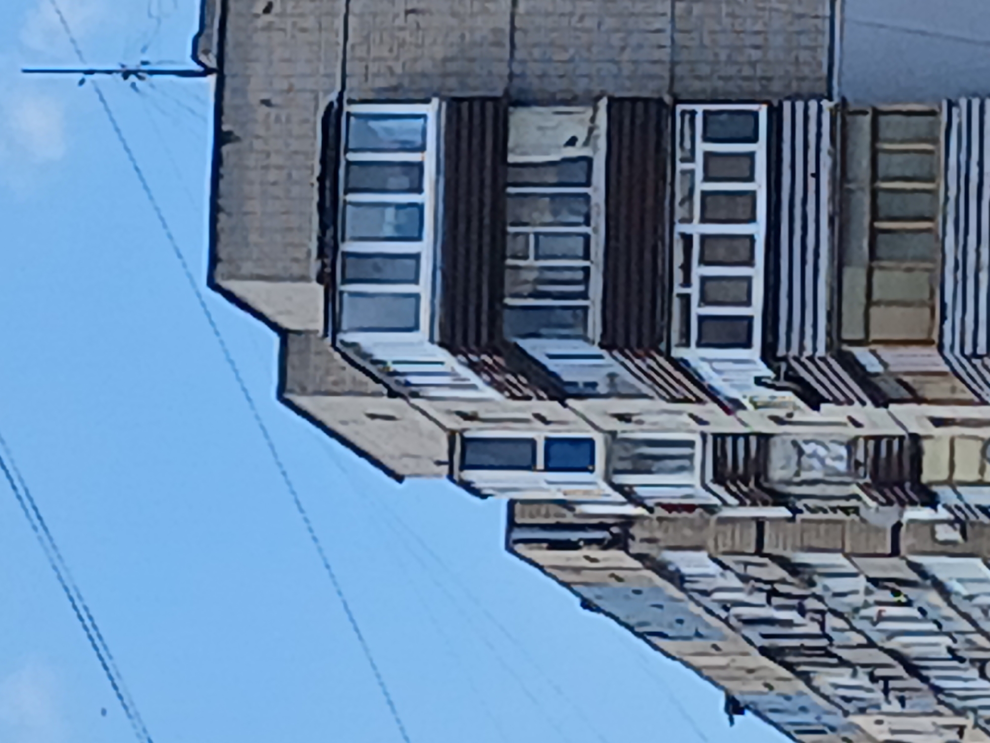 The image depicts the exterior of a multi-story apartment building in Dnipro, Ukraine. The building has a Brutalist architectural style, characterized by its repetitive geometric forms and concrete construction. The facade is a muted gray, with numerous balconies and windows. Some of the balconies are enclosed with glass, while others appear to be open. The windows are a mix of white-framed and some with visible blinds or curtains inside. Power lines are visible crisscrossing the blue sky above the building, with a utility pole on the right side of the frame. The overall impression is of a typical residential building in an Eastern European urban environment.