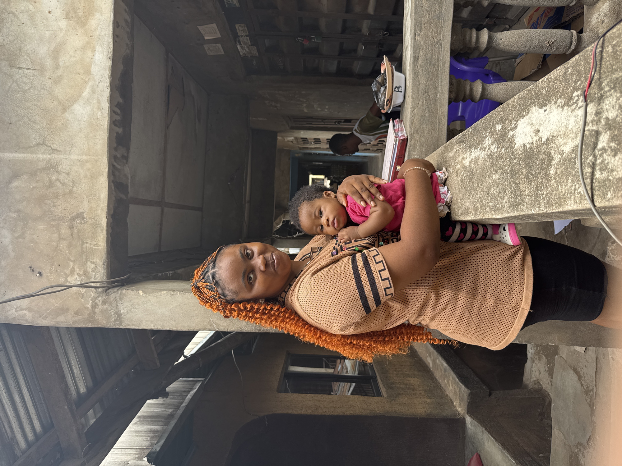 A woman is holding a baby on a concrete balcony overlooking a narrow alleyway in Onitsha, Nigeria. The woman is wearing a tan shirt with black stripes, black shorts, and has orange braided hair. The baby is wearing a pink shirt and patterned tights. In the background, a child is seen sitting on a bench inside a room. The environment is characterized by weathered concrete structures and corrugated metal roofing.