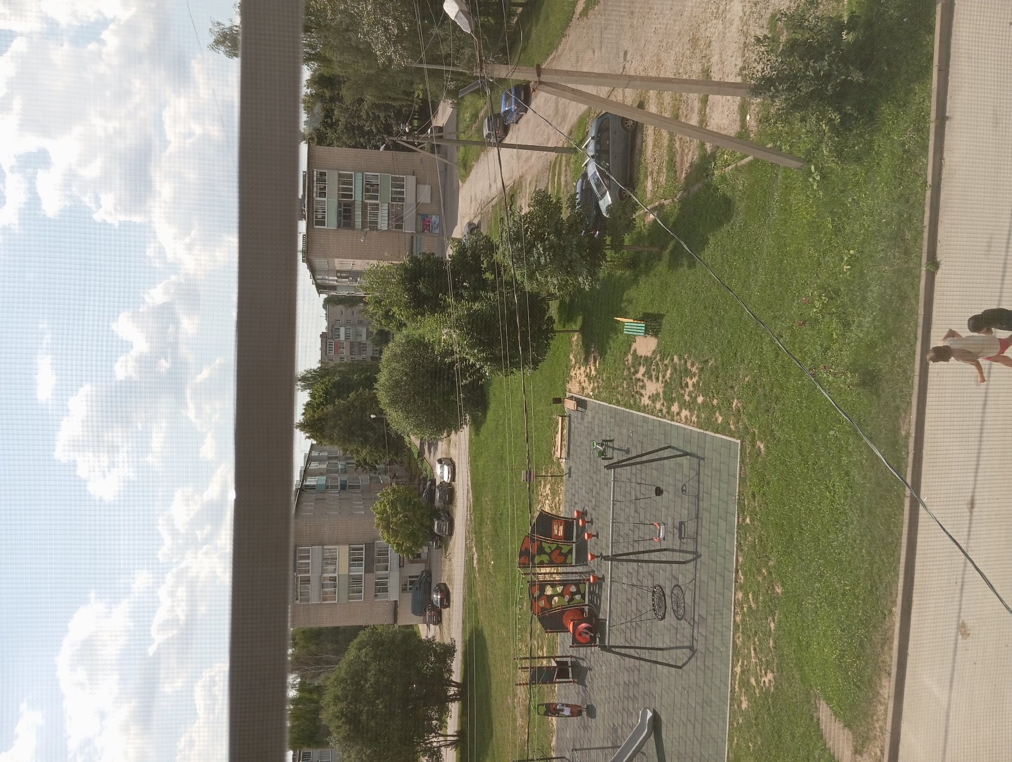 An elevated, eye-level shot captures a residential area with apartment buildings, a playground, and parked cars under a cloudy sky. In the foreground, a children's playground features swings, a slide, and a climbing structure. Beyond the playground, a grassy area is interspersed with trees. Several parked cars are visible on the road and in front of apartment buildings. Two young girls are walking on a paved area in the lower right corner of the image. The scene appears to be in the city of Suvorov, Russia.