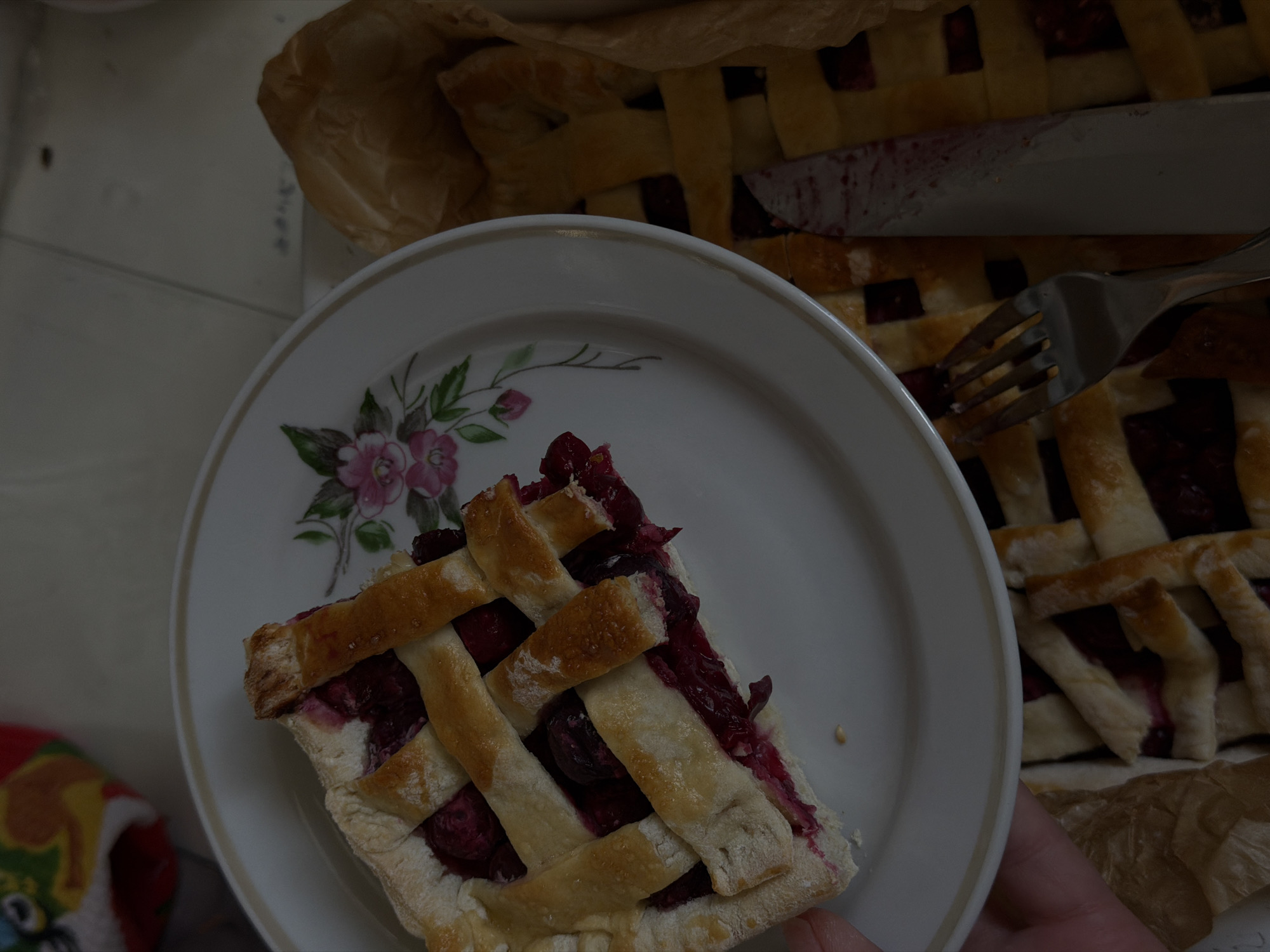A top-down shot shows a slice of cherry pie on a white plate with a floral pattern, adjacent to the whole pie. A fork rests on the pie, and a knife is used to cut it. The scene is likely indoors, with a colorful object visible in the lower left corner.