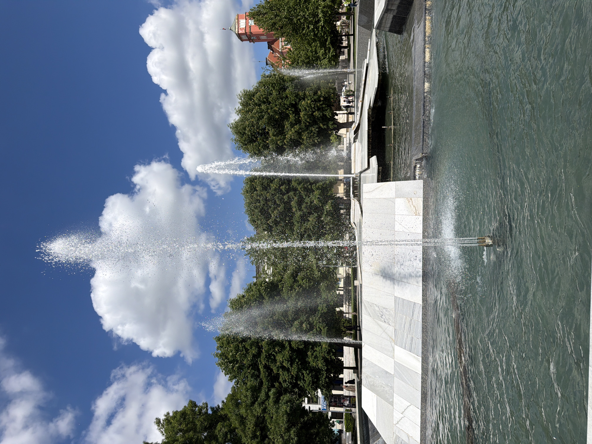The image depicts a water fountain with multiple jets of water shooting upwards. The fountain's structure is made of light-colored stone, possibly marble, with a tiled pattern. Trees surround the fountain. In the background, there is a red brick building with a clock tower. The sky is blue with white, puffy clouds. The location is Pleven, Bulgaria, based on the architectural style of the buildings and the design of the fountain. No people are clearly visible in the main focus of the image, although some may be present in the background. The water from the jets creates a spray effect, capturing the sunlight.