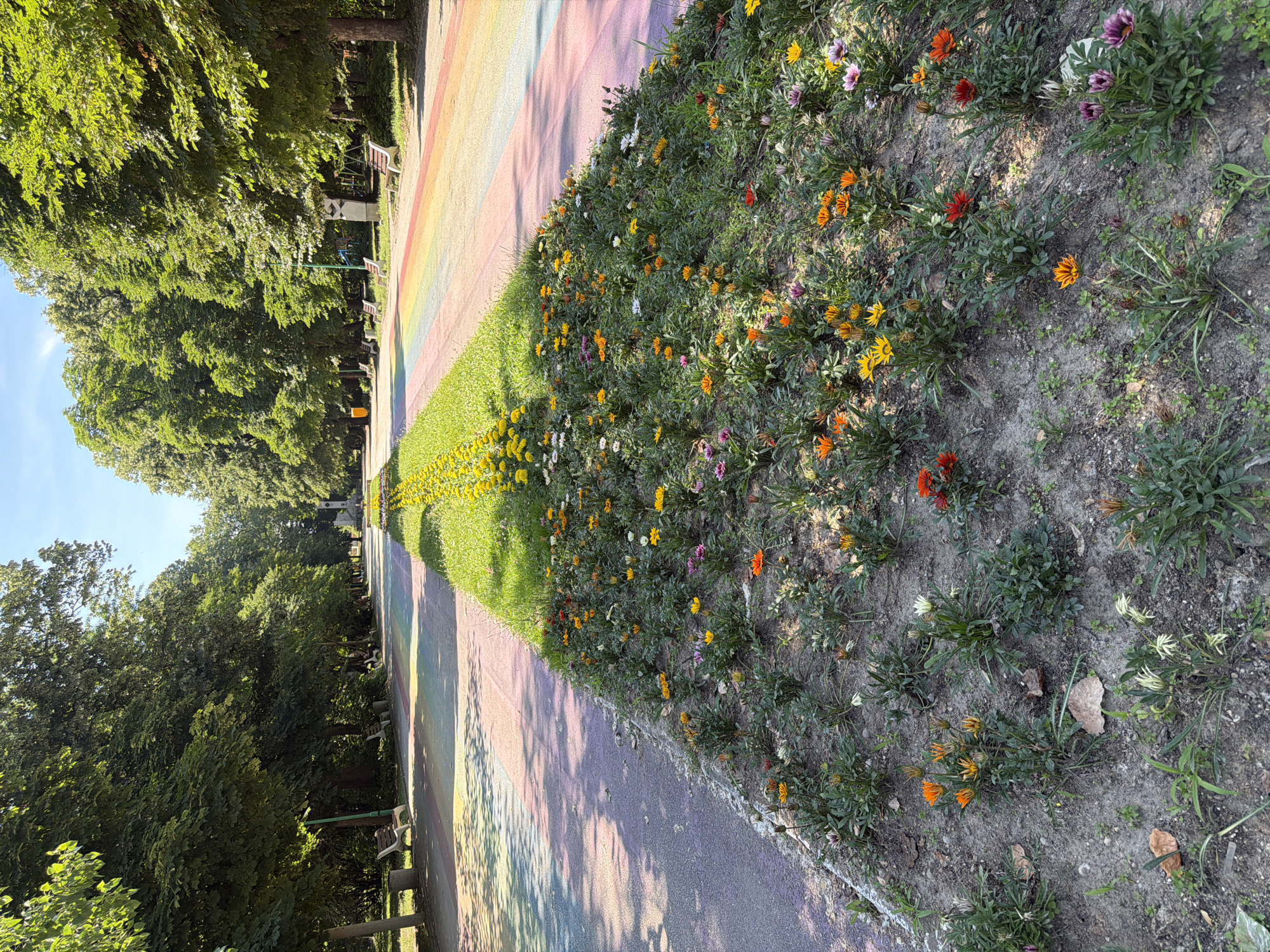 The image depicts a park in Pazardzhik, Bulgaria, featuring a paved pathway adorned with a rainbow-colored design. To the left of the pathway, a flowerbed is densely planted with a variety of colorful flowers, primarily in shades of orange, yellow, white, and purple. The flowers are in bloom and appear healthy, with lush green foliage. Beyond the flowerbed, mature trees line the pathway, creating a canopy of shade. On the right side of the pathway, another flowerbed or grassy area is visible, also featuring flowering plants. In the background, more trees and a clear blue sky suggest a sunny day. Benches are visible along the sides of the pathway, indicating a public space designed for relaxation and recreation. The overall scene conveys a vibrant and well-maintained park environment.