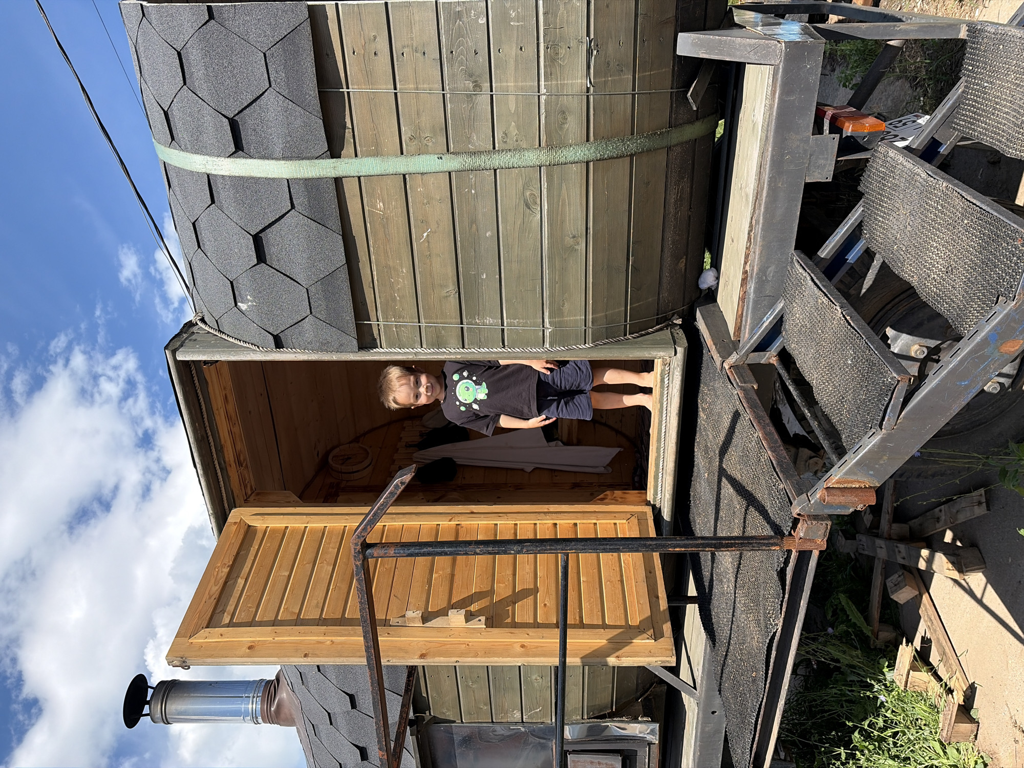 The image shows a young boy standing inside a wooden sauna mounted on a trailer. The boy is wearing a black t-shirt with a green alien-like graphic and dark shorts. He is looking towards the camera with a slight smile. The sauna has a rounded wooden exterior covered with dark grey hexagonal shingles. A green strap is visible around the sauna's midsection, securing it. The trailer structure is made of metal and appears somewhat rusted, with parts of the trailer and what looks like a spare tire visible.
The scene is set outdoors under a bright blue sky with scattered white clouds, suggesting daytime, likely sunny weather. The ground appears to be a mix of dirt and gravel, with some green grass and other vegetation growing around the trailer. A metal chimney pipe extends from the sauna's roof. The overall impression is of a rustic, possibly mobile sauna setup in a rural or backyard setting. The image has been rotated for display, with the sky appearing at the top left.
