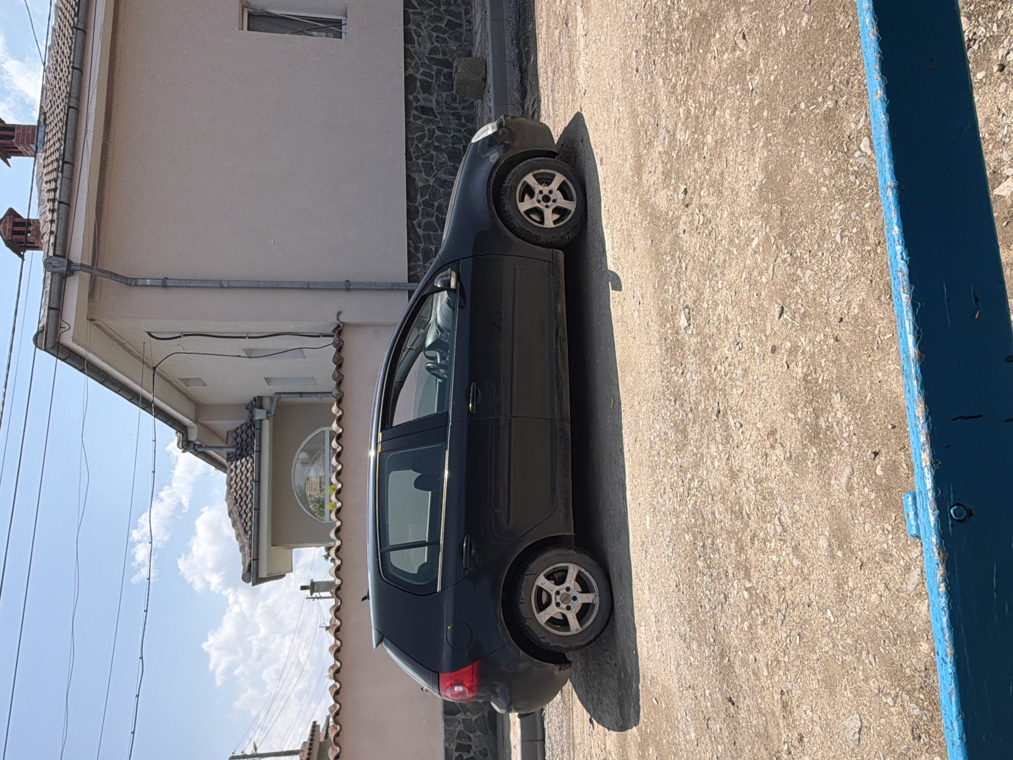 An outdoor, rotated full-shot photograph captures a parked dark-colored five-door hatchback car, likely a Volkswagen Golf Mk5, in what appears to be a residential area in Dabene, Bulgaria. The car is positioned on an unpaved, gravelly or dirt surface, casting a long shadow to its left, indicating bright mid-day to afternoon sunlight. Its alloy wheels appear somewhat dusty.
In the background, a light-colored building with a rectangular window is visible, possibly a house. To the car's right, the lower section of this building features a patterned stone facade. Further back and to the left, another building with a brown-tiled roof and distinctive rounded window can be seen. Utility lines crisscross the clear blue sky, which has scattered white clouds. Rain gutters and downspouts are visible along the edges of the buildings, and two chimney-like structures cap one of the roofs.
On the far right of the frame, a weathered blue wooden fence post or part of a gate stands prominently, showing chipped paint and a rough texture. The overall setting suggests a sunny day in a quiet, perhaps rural or semi-urban, residential environment. No people are visible, and no specific activity is taking place other than the car being parked. There is no visible text in the image.