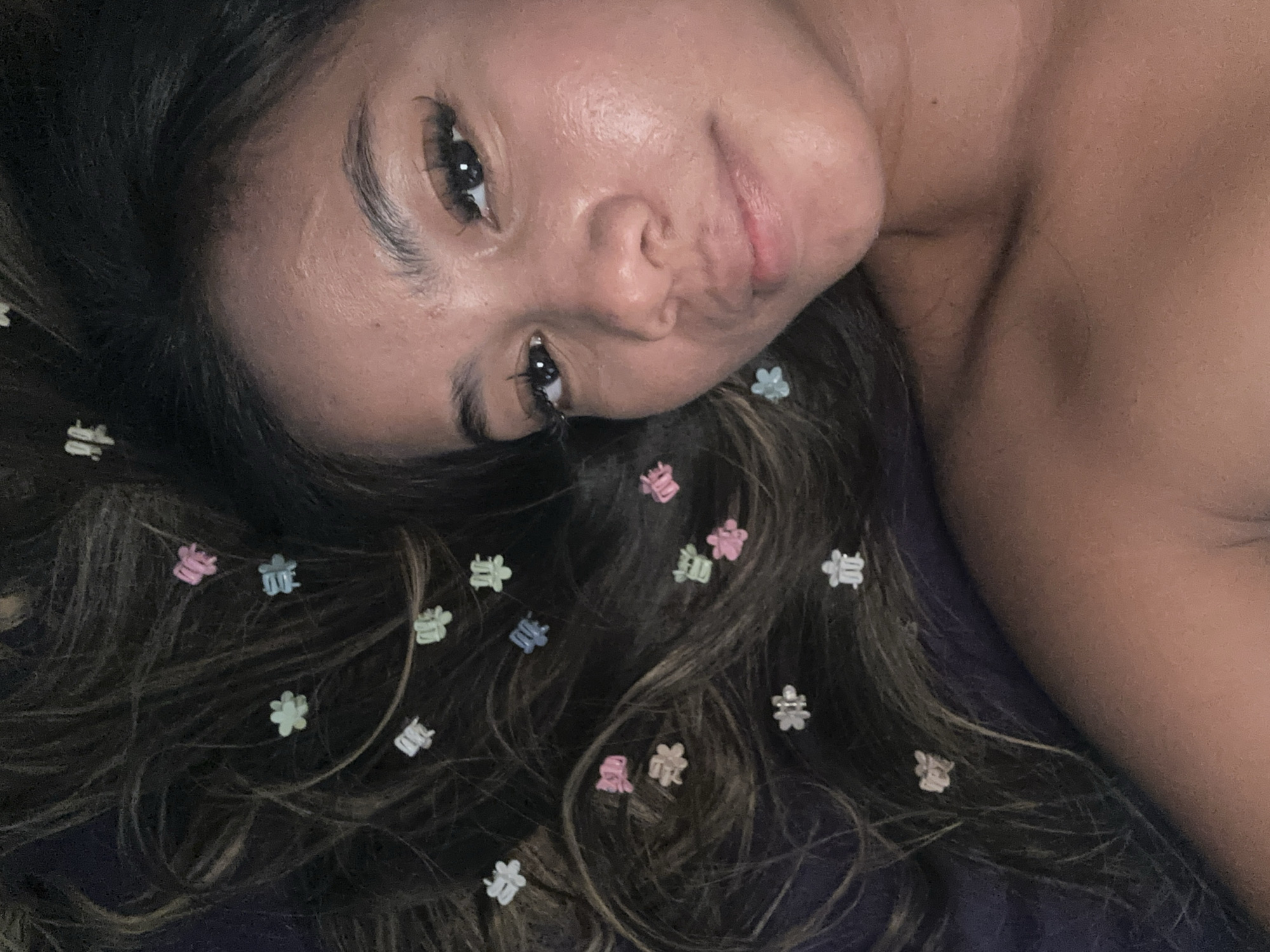 A young woman with long, dark hair lies on a dark-colored surface. Her hair is adorned with numerous small, colorful flower-shaped hair clips. She is facing the camera with a slight smile. The image is a close-up shot. There is no discernible evidence to indicate that this image was taken in North Jakarta, Indonesia.