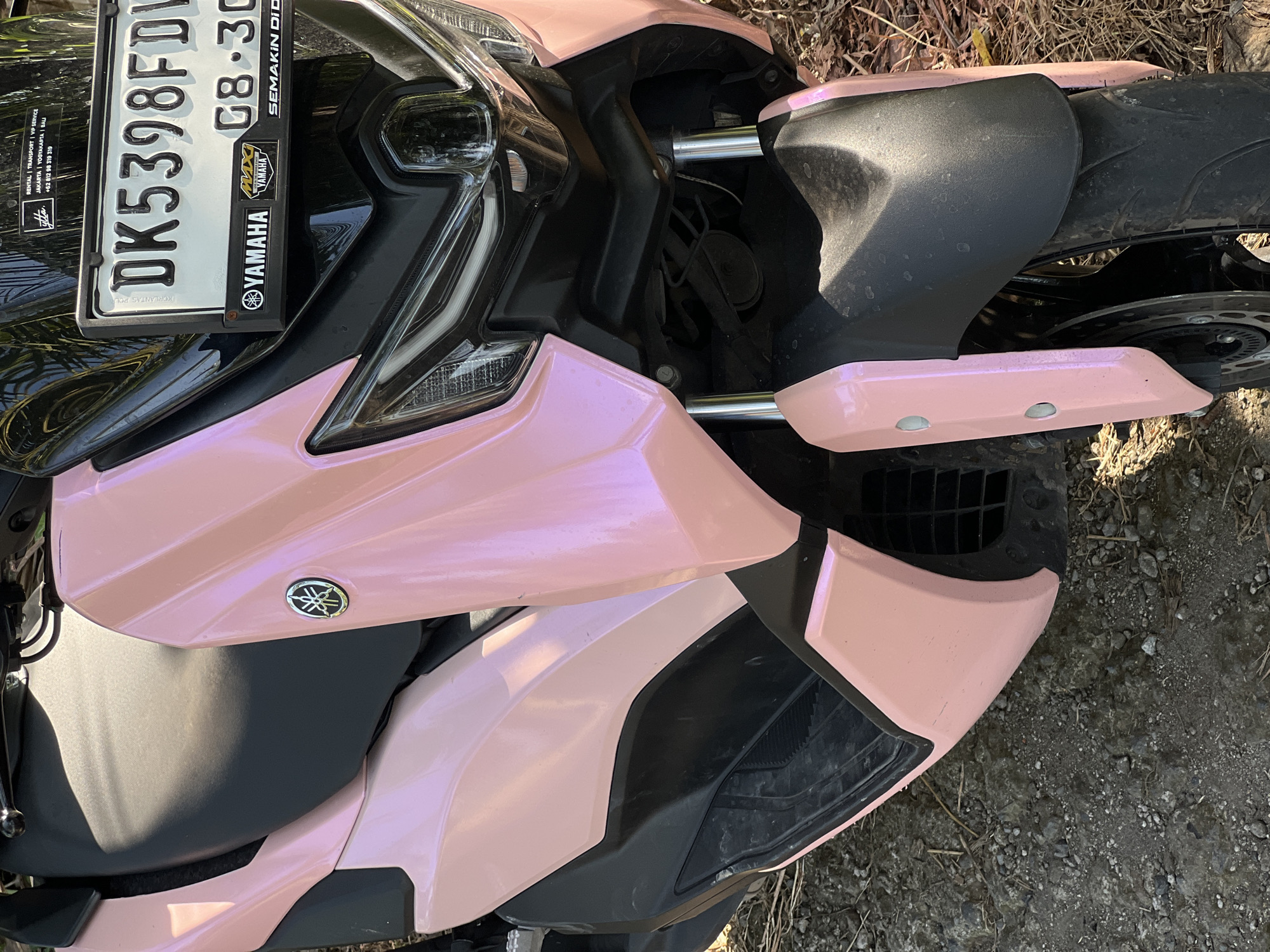 A close-up, high-angle shot shows the front of a pink motorcycle parked on a dirt and gravel surface, suggesting a rural or unpaved area, possibly in or near Kerambitan, Indonesia. The motorcycle has a black seat and black accents on the body. A license plate with the characters "DK5398FDW" and "08-30" is visible on the front. Yamaha logos are present on the motorcycle and on stickers below the license plate. The front wheel and fender are visible, as are parts of the headlight assembly. The scene is outdoors, with dry grass and twigs on the ground, indicating a sunny day.