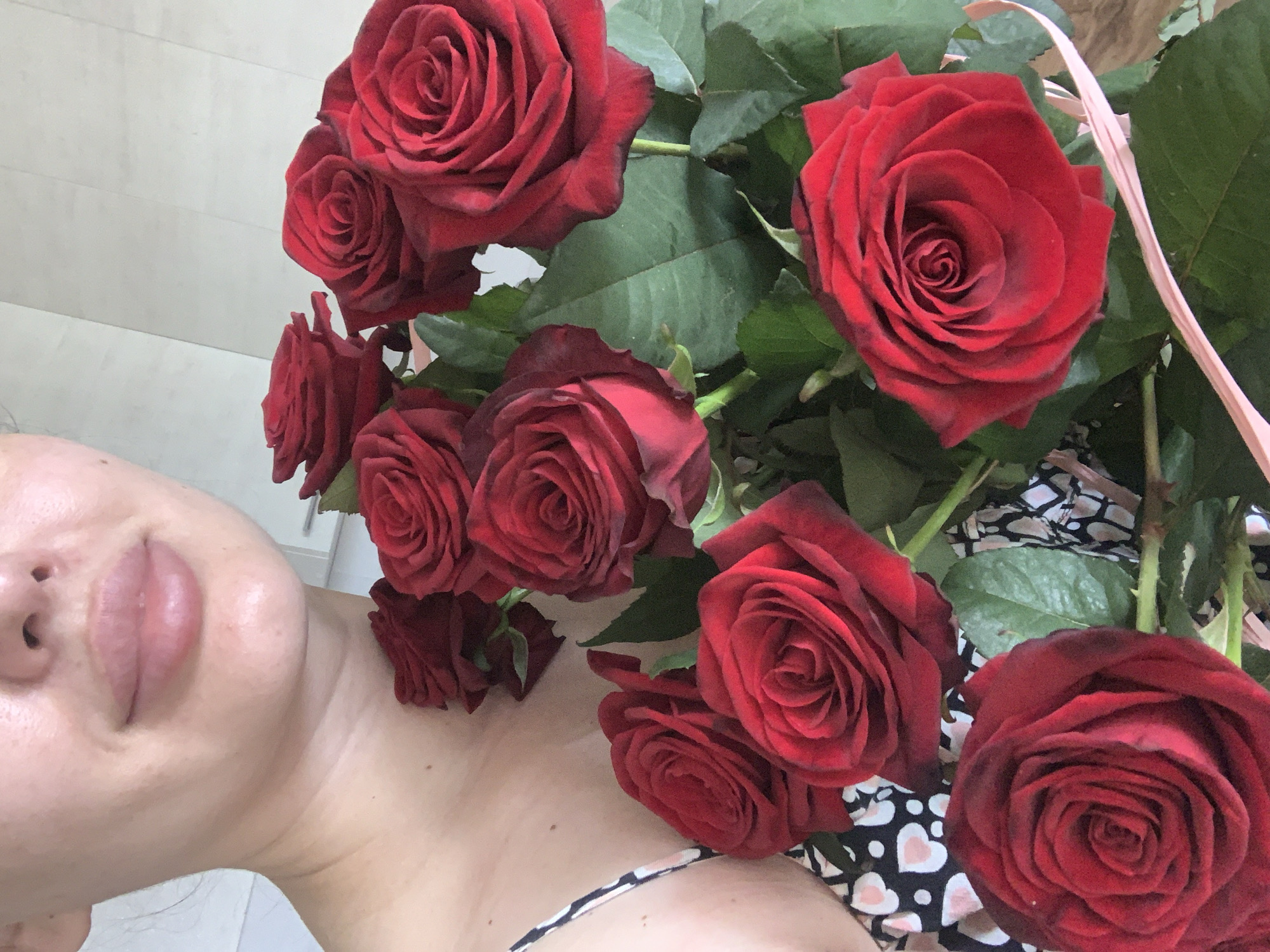 A close-up shot features a woman with fair skin, full lips, and a visible nose, positioned on the left side of the frame. A bouquet of deep red roses occupies the right side and extends over the woman's shoulder and chest. The roses are in various stages of bloom, and some green leaves are visible amongst the flowers. The woman is wearing a patterned top with hearts. The background appears to be a light-colored wall. The location of Ternopil, Ukraine, cannot be confirmed based solely on visual information.
