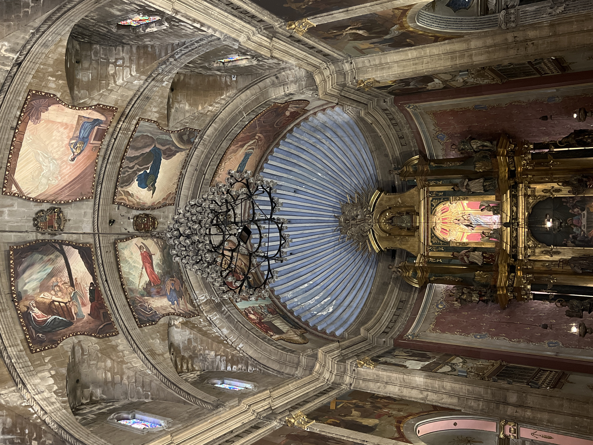 The image depicts the interior of a grand church, likely a cathedral or a significant parish church, with an ornate altar at the center. The ceiling is adorned with intricate frescoes depicting religious scenes and figures, framed by stone arches. A large, multi-tiered chandelier hangs from the center of the ceiling, casting a warm light on the scene. The walls are also decorated with large paintings and religious iconography. The altar itself is a complex structure, featuring multiple statues and gilded ornamentation. The overall impression is one of historical and religious grandeur, with a focus on elaborate artwork and architecture. The location is the Church of Our Lady of the Angels in Pollença, Spain.
