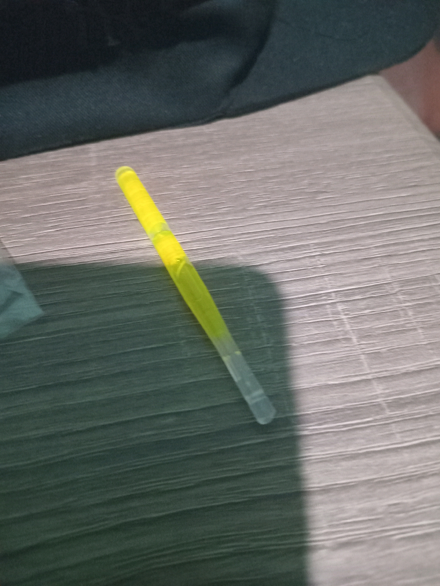 A fluorescent yellow glow stick is positioned diagonally on a textured, light-colored surface. The glow stick appears to be activated, emitting a bright yellow light, with a portion of it transitioning to a translucent white at its lower end. A dark shadow is cast by the glow stick onto the surface. In the upper portion of the image, a dark, soft fabric is visible. The scene is likely indoors, possibly on a table or countertop, and there is no visible indication of the city of Junjung, Indonesia. The image depicts a static object without any discernible human presence or activity.