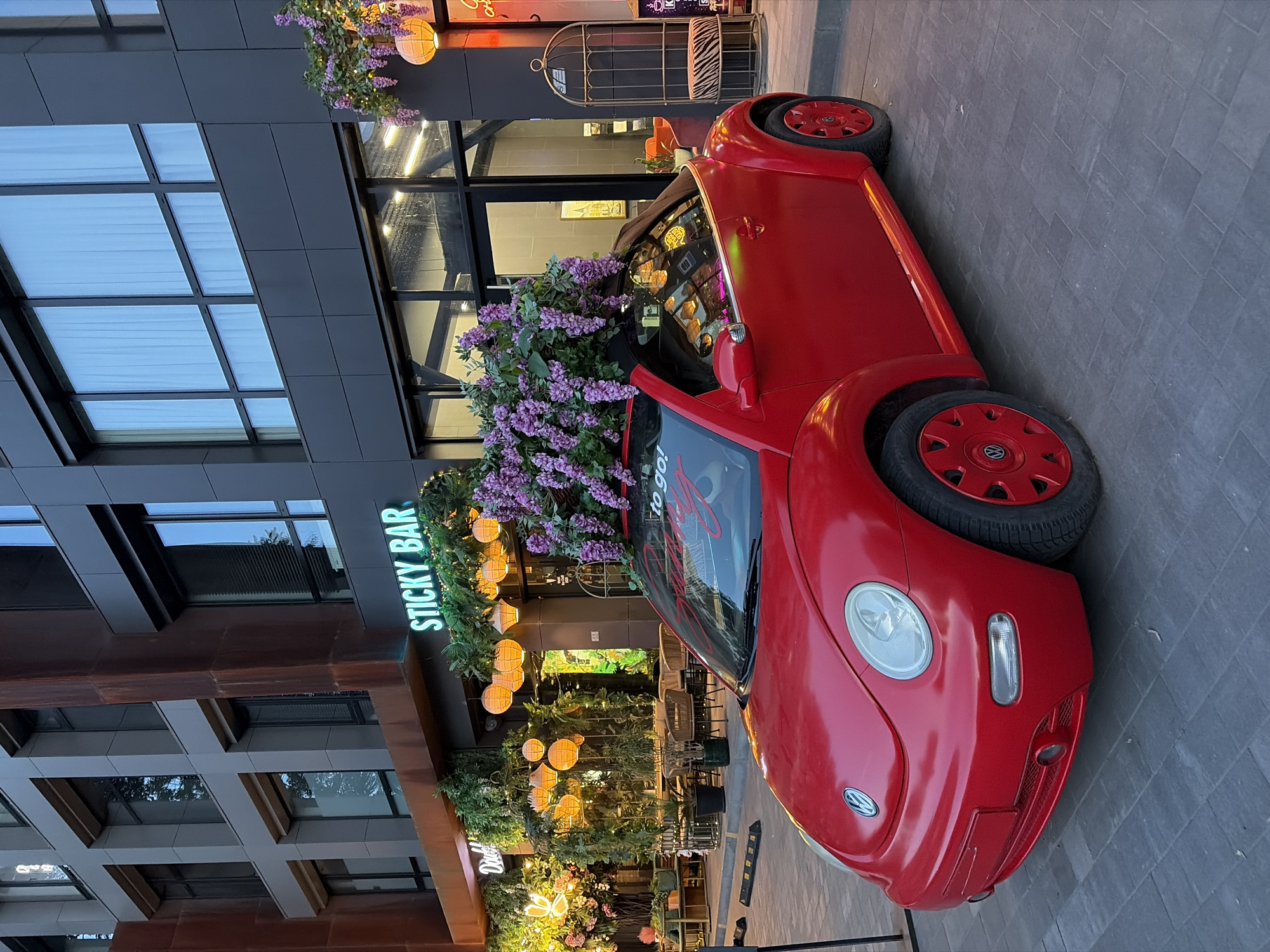 The image depicts a vibrant scene outside a establishment in Dnipro, Ukraine. In the foreground, a bright red Volkswagen Beetle convertible is parked, its top down and adorned with an abundant arrangement of purple flowers cascading over the windshield and hood. The car's license plate is not visible.
Behind the car, the facade of a modern building is prominent, featuring dark gray panels and large windows. To the left, a sign with green neon lettering reads "STICKY BAR." The building's ground floor appears to be a restaurant or cafe with outdoor seating visible through a glass storefront. Lush green foliage and string lights with spherical lanterns are used as decorative elements, creating a welcoming ambiance.
The sidewalk is paved with gray rectangular tiles. The overall impression is one of a stylish and inviting urban setting, likely a popular spot for dining and socializing. The time of day appears to be late afternoon or early evening, as suggested by the warm lighting from the lanterns and the soft glow illuminating the building's interior.