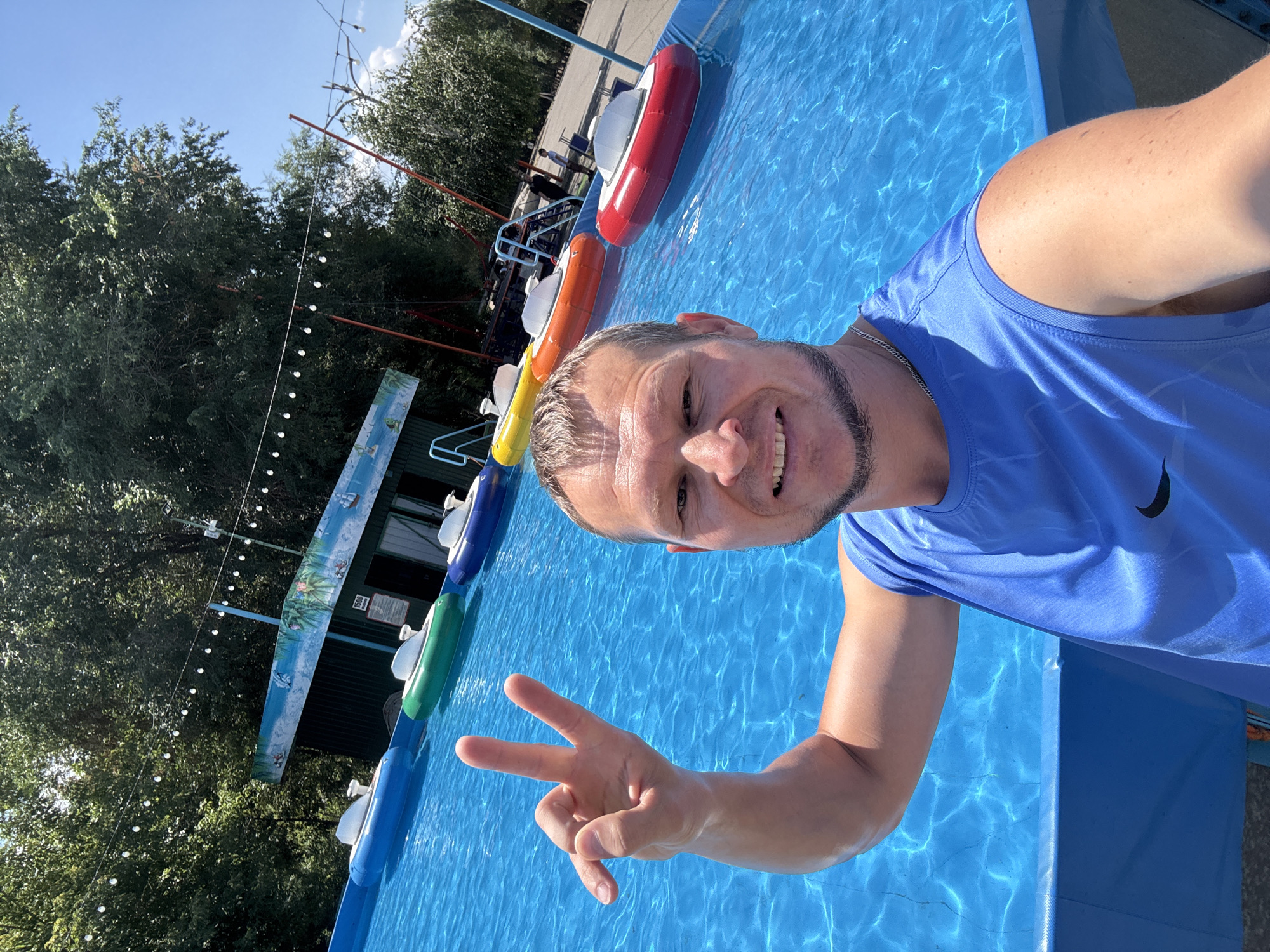 The image shows a man taking a selfie in an outdoor pool. The man is wearing a blue tank top and has a beard. He is smiling and making a peace sign with his left hand. In the background, there are several inflatable boats in the pool, and beyond that, trees and a building. The pool appears to be part of an amusement park or recreational area. The location is stated to be the city of Oskemen, Kazakhstan. The weather is sunny, and the sky is clear blue. The water in the pool is a vibrant blue color, and the sunlight is reflecting off the surface. The overall atmosphere of the image is cheerful and relaxed.