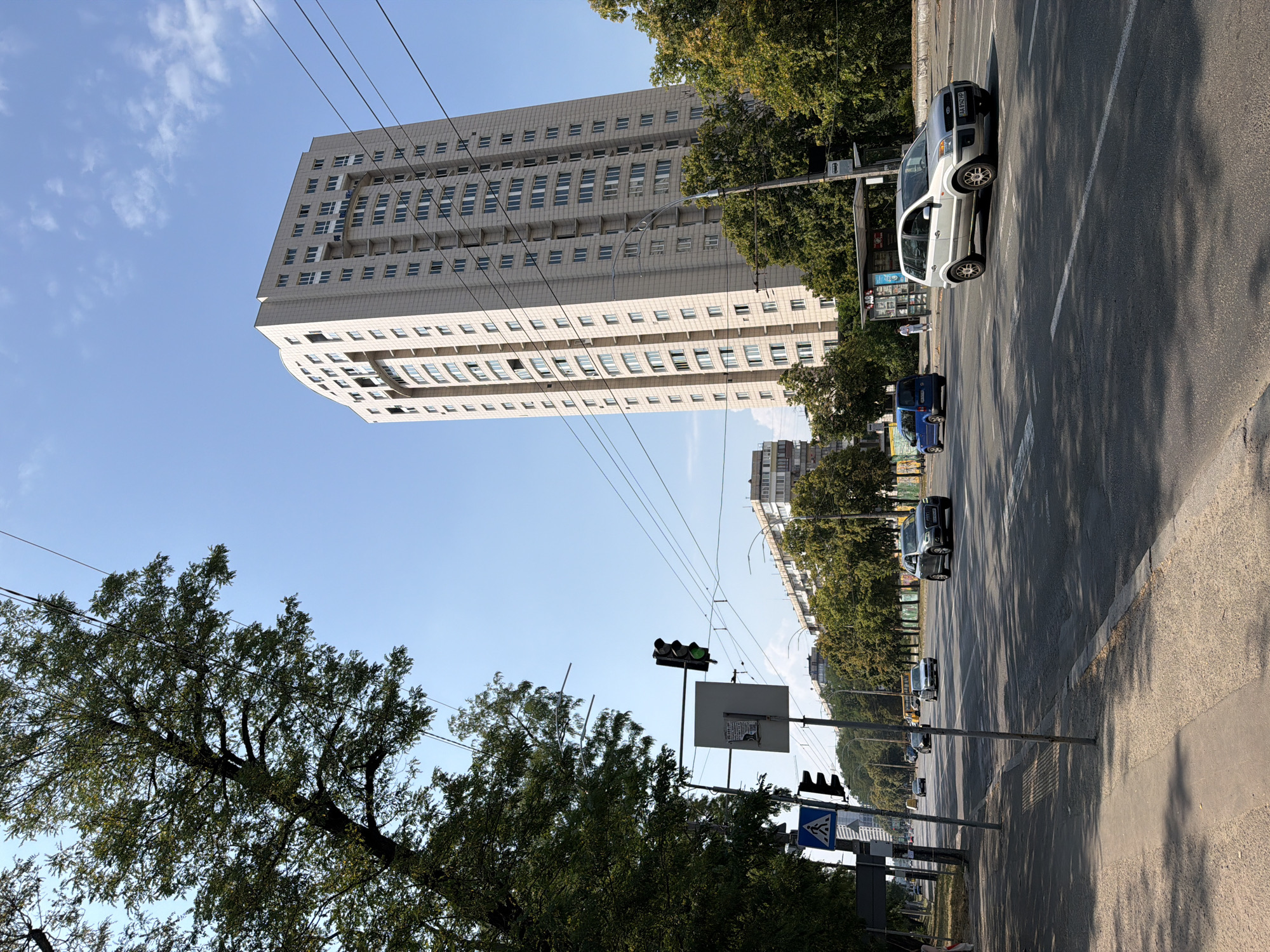 The image depicts a street scene in Kyiv, Ukraine, on a clear, sunny day. A tall, modern apartment building dominates the skyline on the right side of the frame, characterized by its light-colored facade and numerous windows. In the foreground, a multi-lane asphalt road is visible with several cars driving on it. A pedestrian crossing is marked on the road, and a traffic light is illuminated green. On the left side of the street, mature trees line the sidewalk, casting shadows on the road. Further down the street, other buildings and more trees can be seen. A bus stop shelter is present on the right side of the road. The overall atmosphere is one of urban activity under a bright blue sky with sparse clouds.