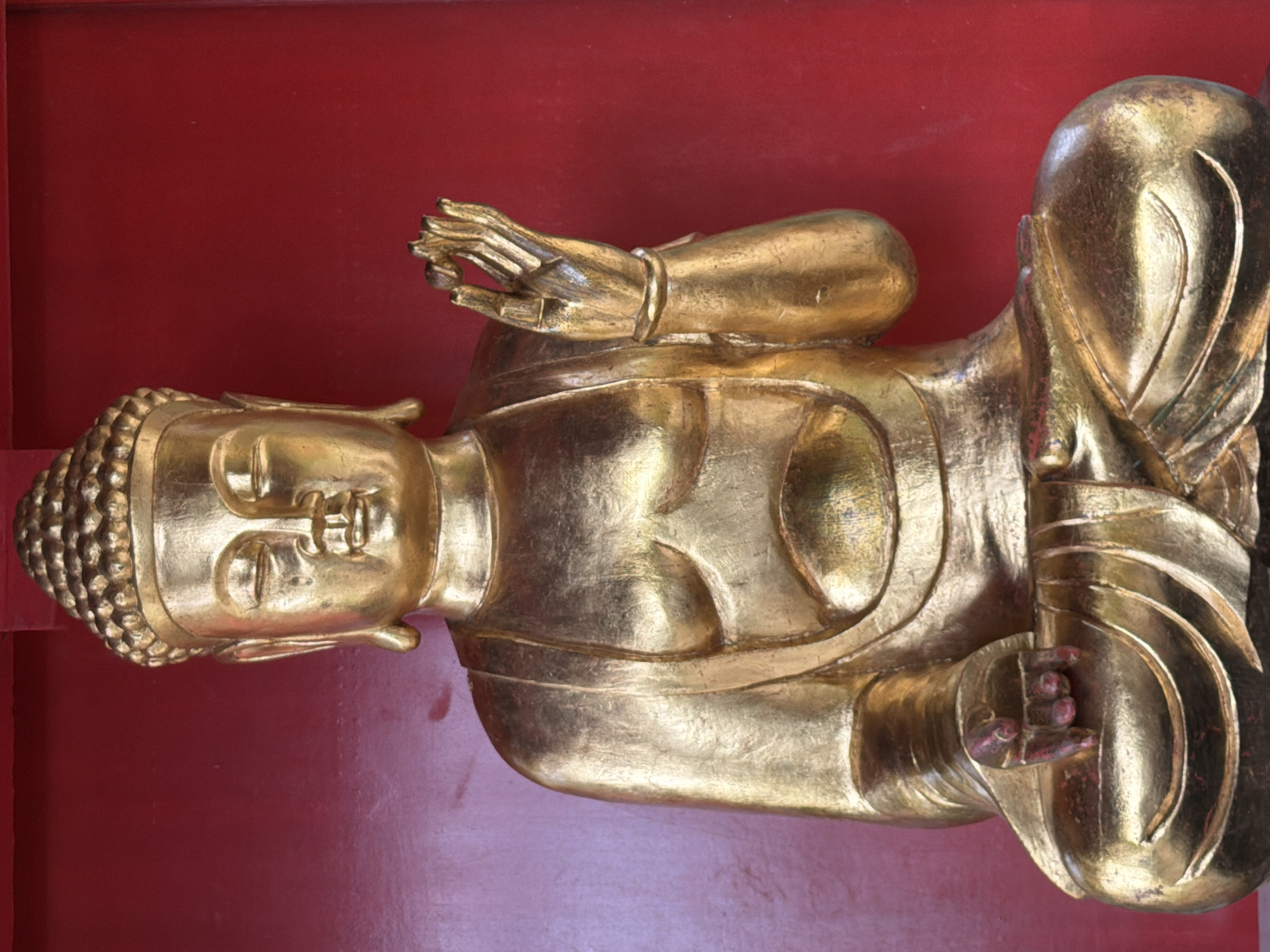 A full-shot, eye-level, front-facing studio photograph shows a golden statue of the Buddha. The statue is seated in a cross-legged position, with its right hand raised and fingers in a mudra, while its left hand rests on its lap. The Buddha statue is positioned against a plain, bright red background, with a shallow recess behind it. The statue appears to be made of polished gold, reflecting light and showing subtle variations in color due to the texture. The facial features are serene, with closed eyes and a slight smile. The robes are draped over the left shoulder, and the chest is bare. The statue is detailed and appears to be a traditional depiction of the Buddha. The image does not contain any elements that would indicate a specific location in Funchal, Portugal.